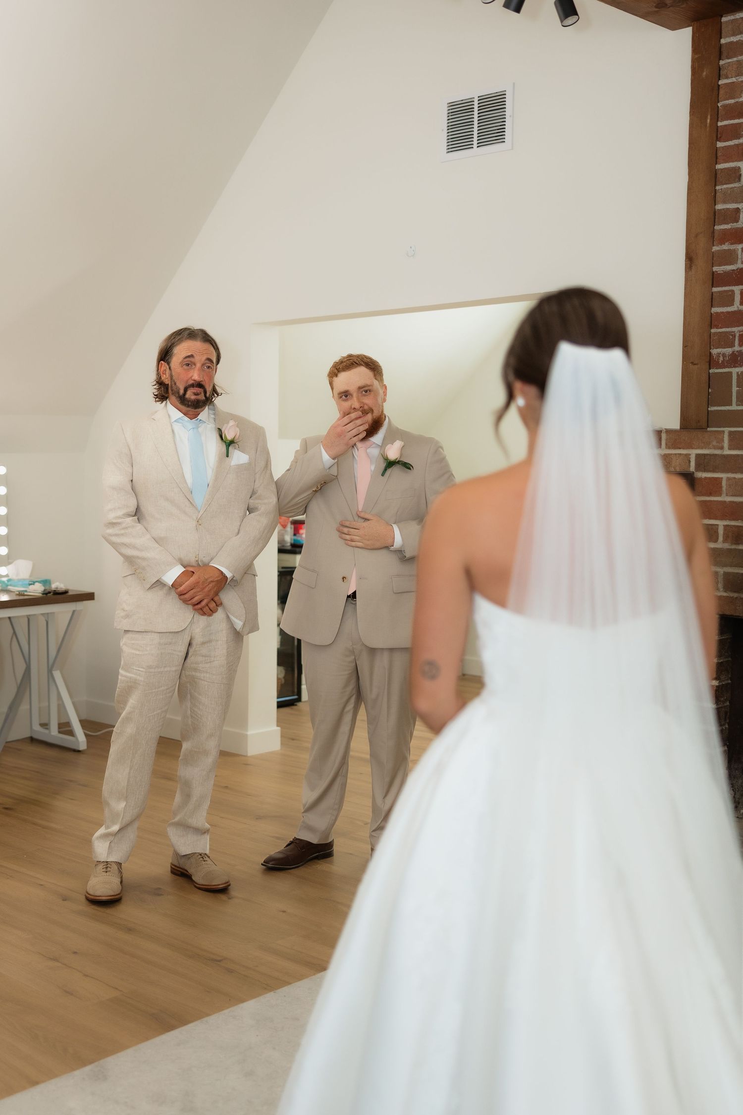 Groom reacts with surprise and emotion as bride reveals her wedding dress during a first look moment indoors.