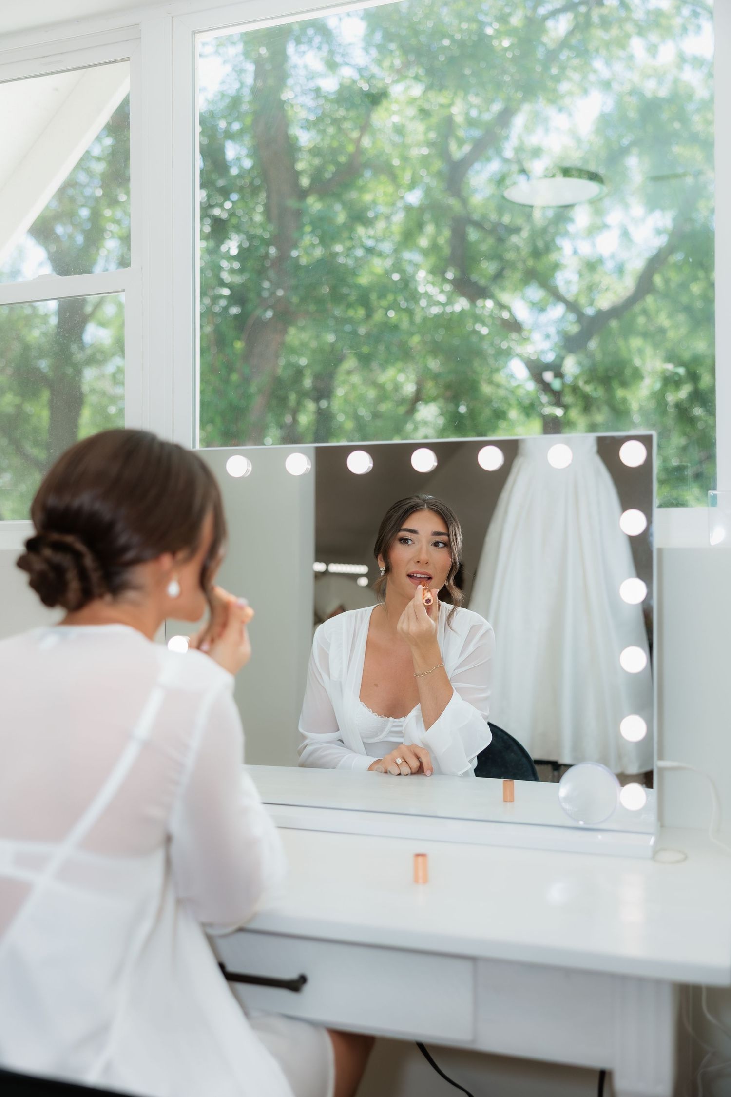 Bride applying lipstick at vanity mirror with Hollywood lights during wedding preparations.