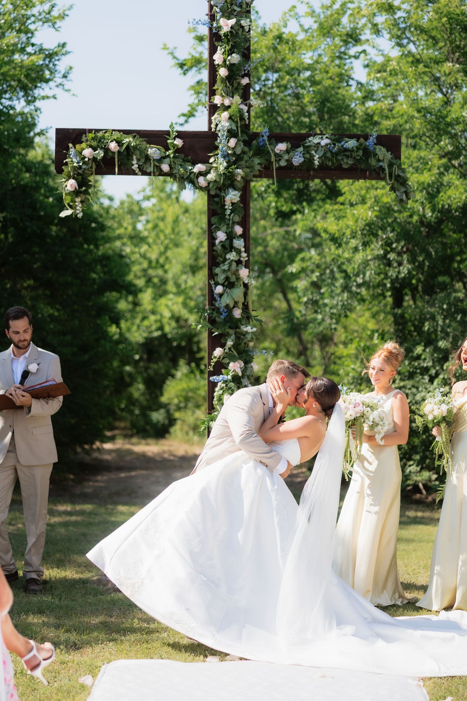 Bride and groom share first kiss beneath floral-adorned wooden cross at outdoor wedding ceremony.
