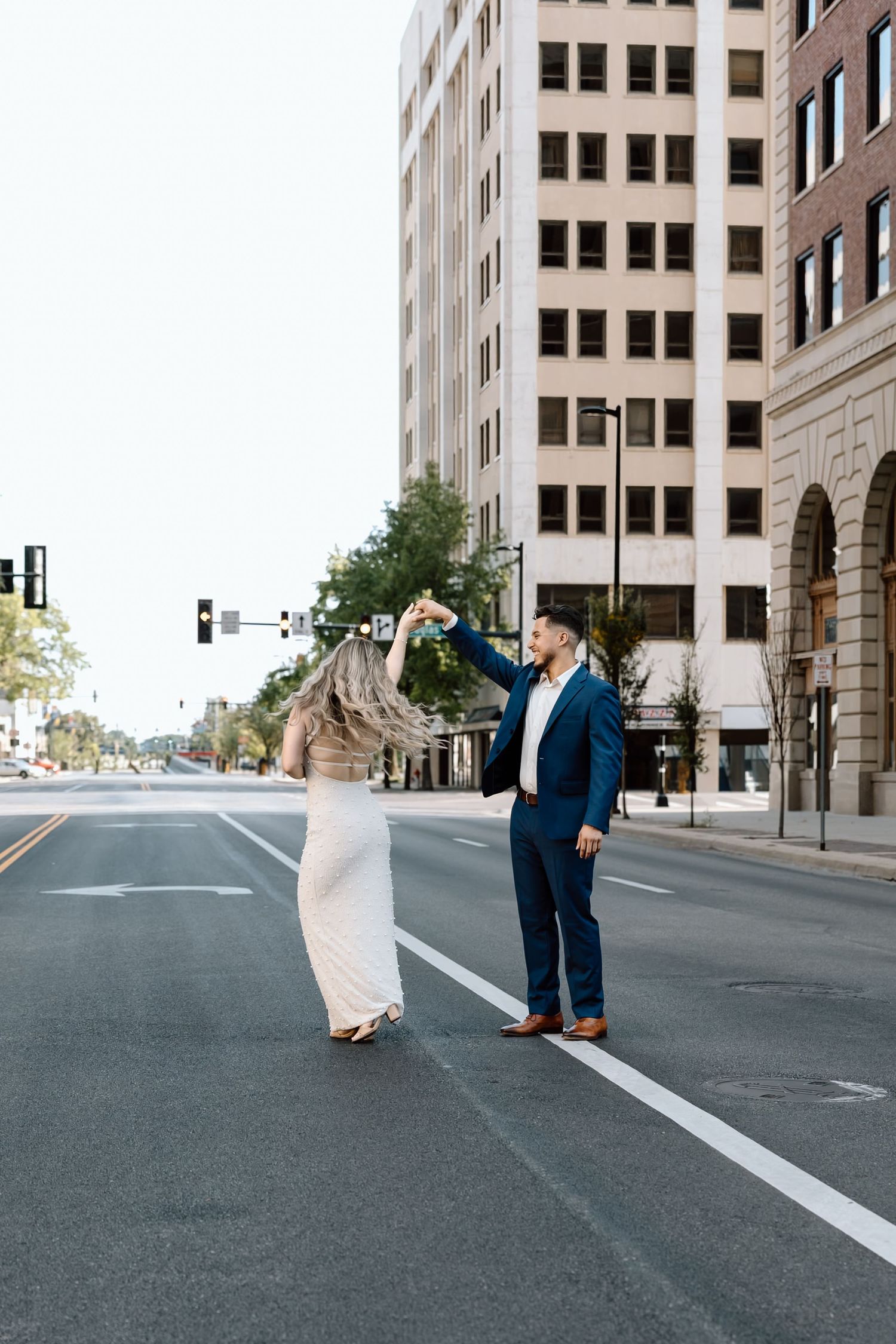 Couple dancing in empty city street, man in navy suit spinning woman in white sequin dress.