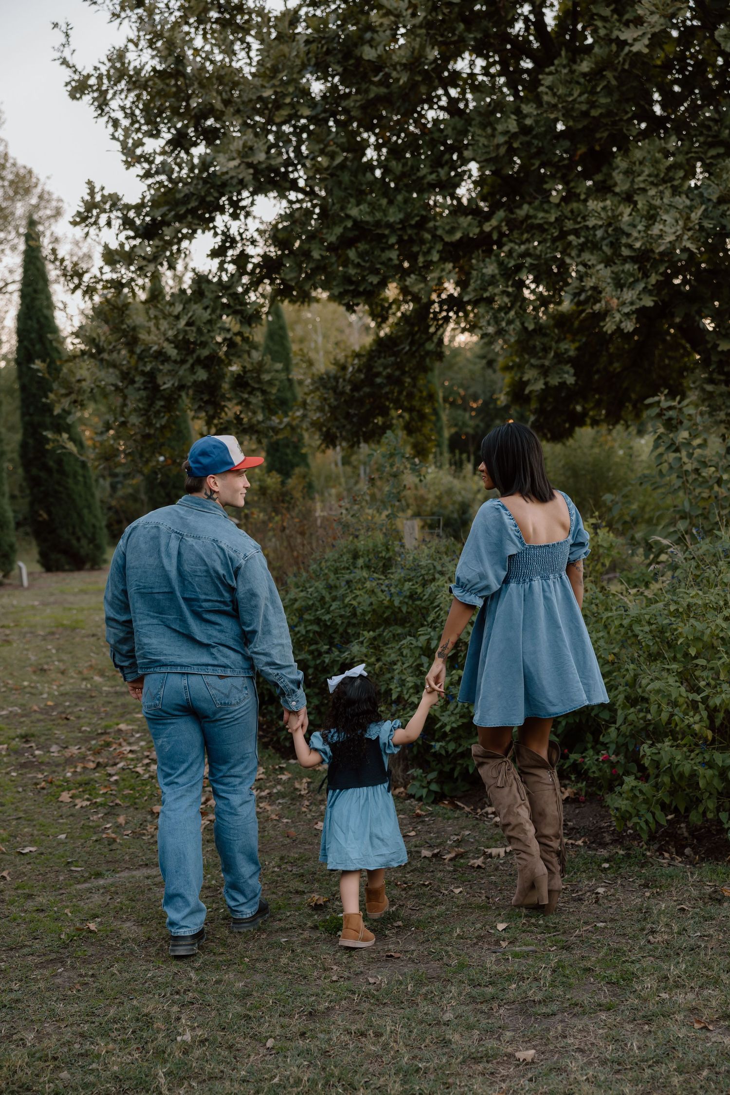 A family of three in coordinating denim outfits walks hand in hand through a lush green garden path.