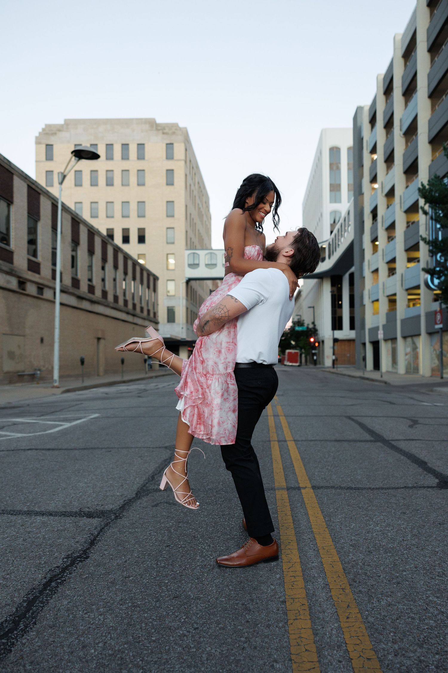Man lifting woman in a pink dress on an urban street, sharing a joyful romantic moment at dusk.