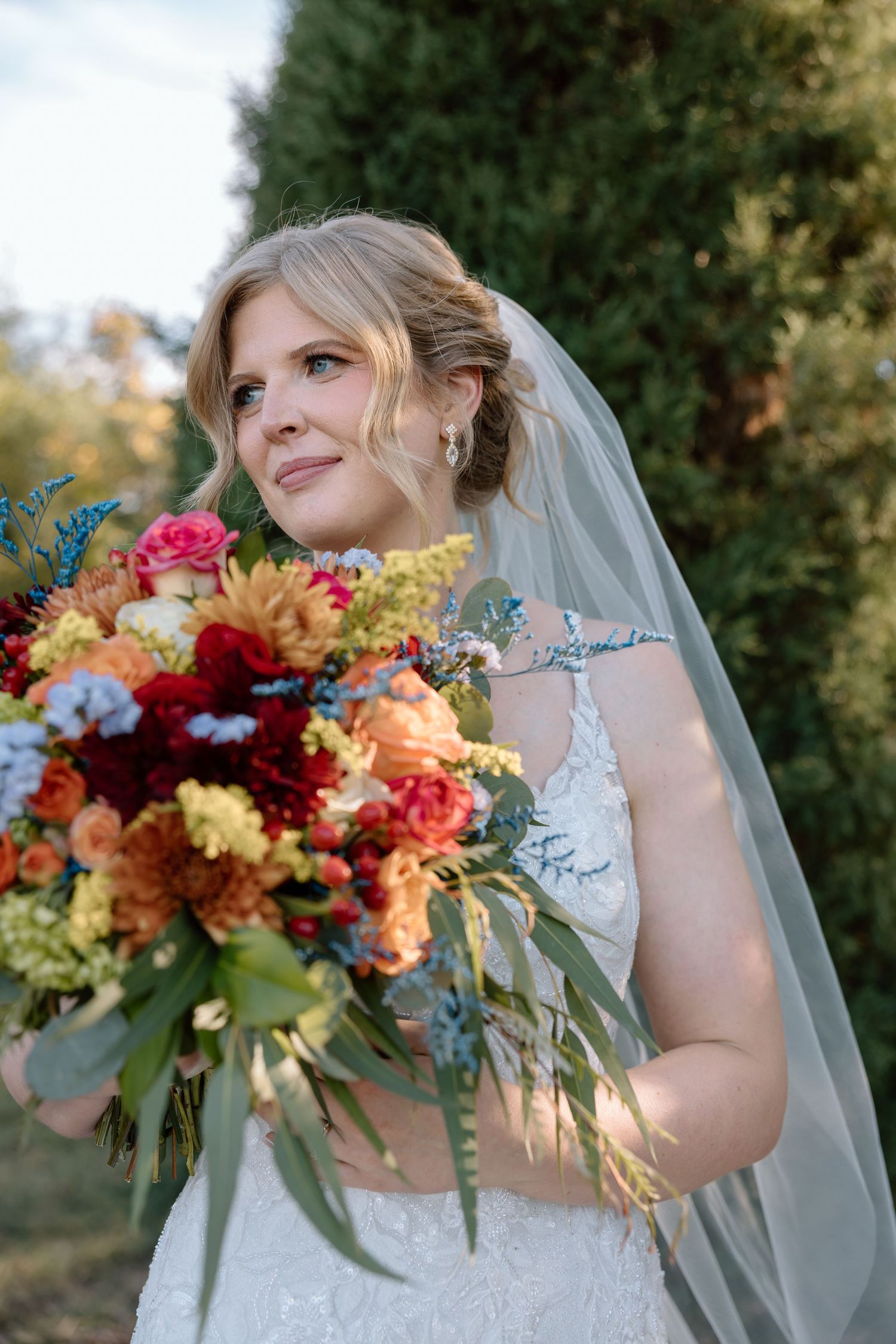 Beautiful blonde bride in lace wedding gown holding a vibrant colorful bouquet with veil flowing outdoors.