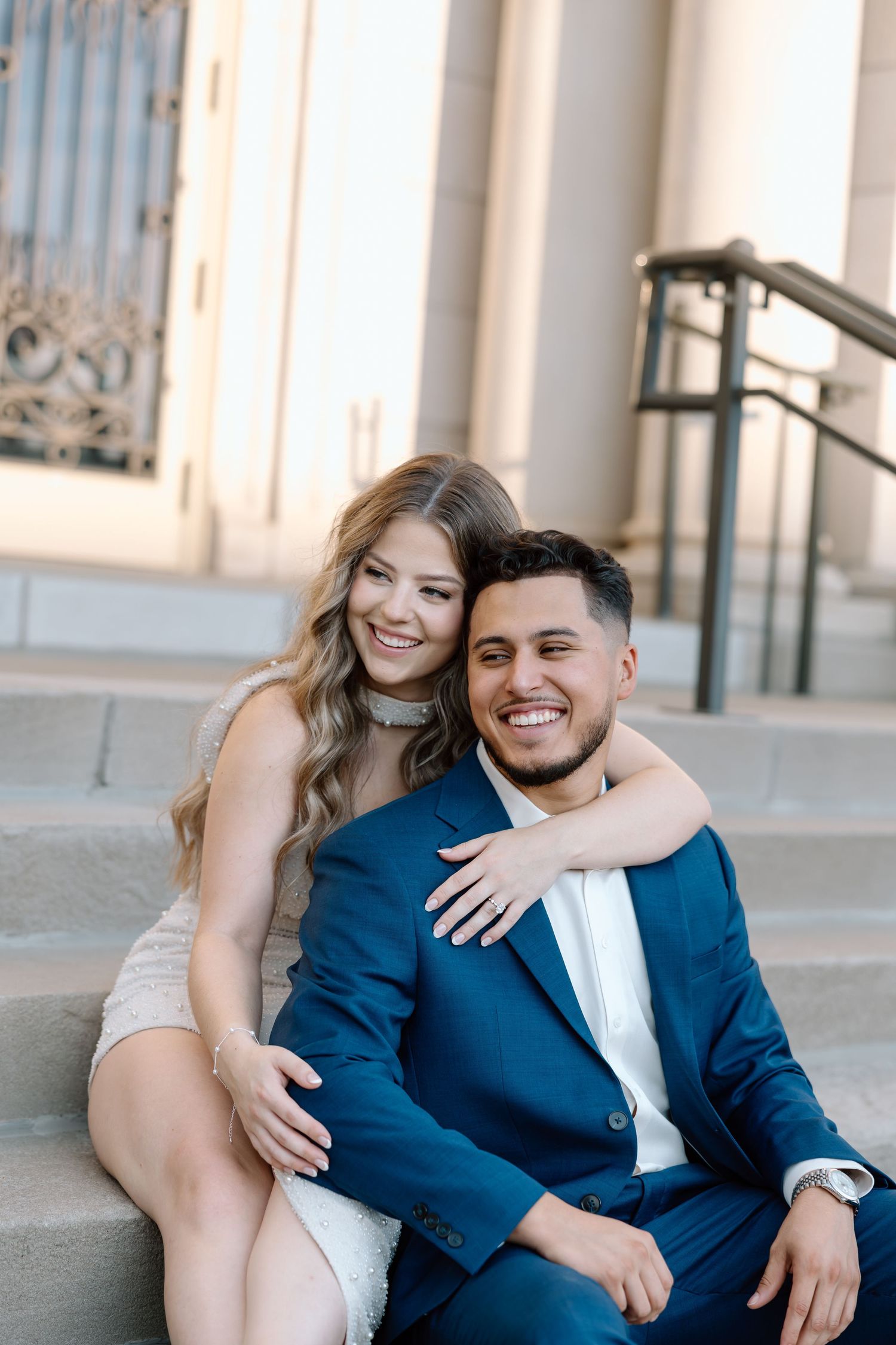 Smiling couple in formal attire sitting on stone steps, woman in white dress embracing man in navy suit.