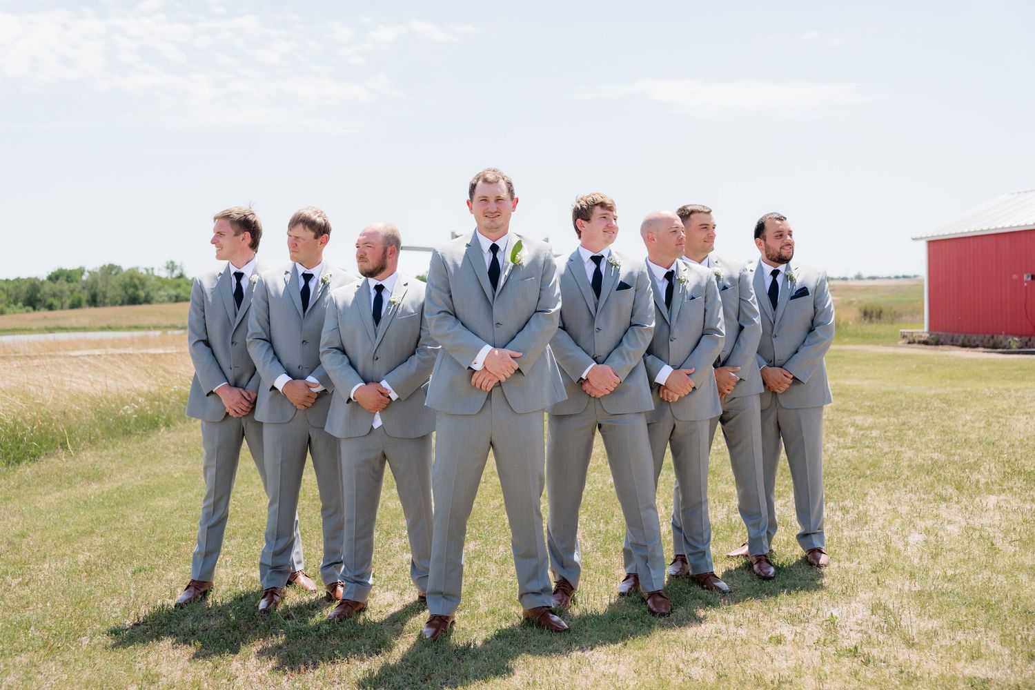 Groom and groomsmen in matching gray suits and black ties pose in a line on a sunny green field.