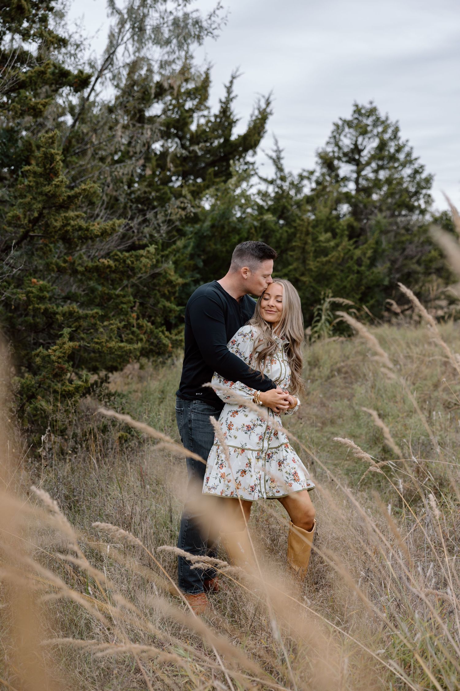 Man kissing woman in a field of tall golden grass with pine trees behind them.