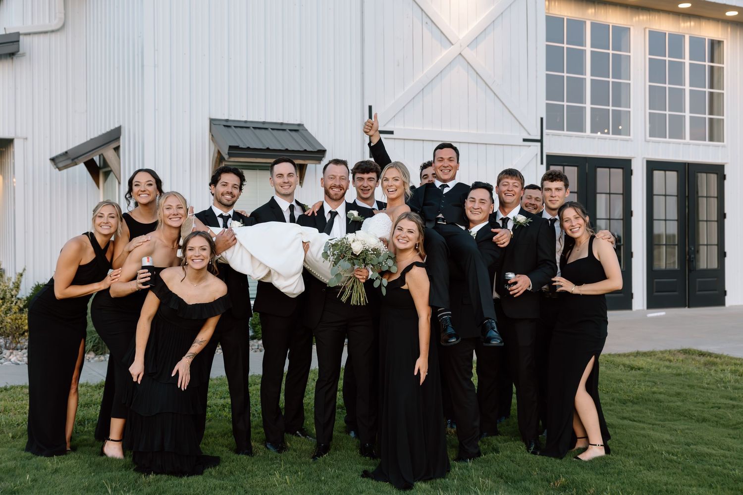 Wedding party in black attire poses joyfully outside a white barn venue, with bride being lifted by groomsmen.