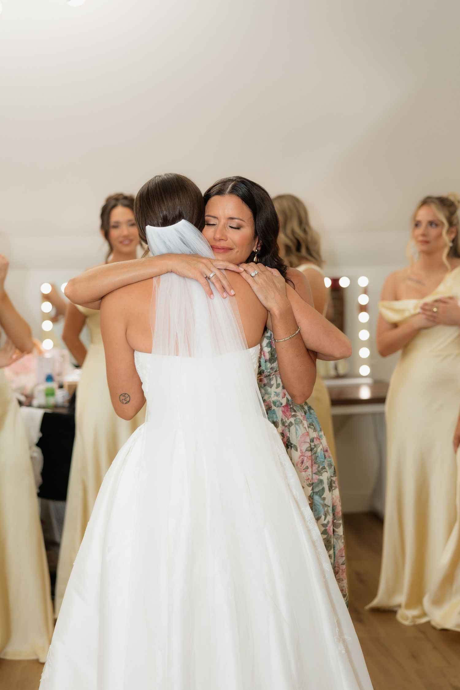Emotional bride and bridesmaid share a heartfelt hug in a bridal suite surrounded by bridesmaids in champagne gowns.