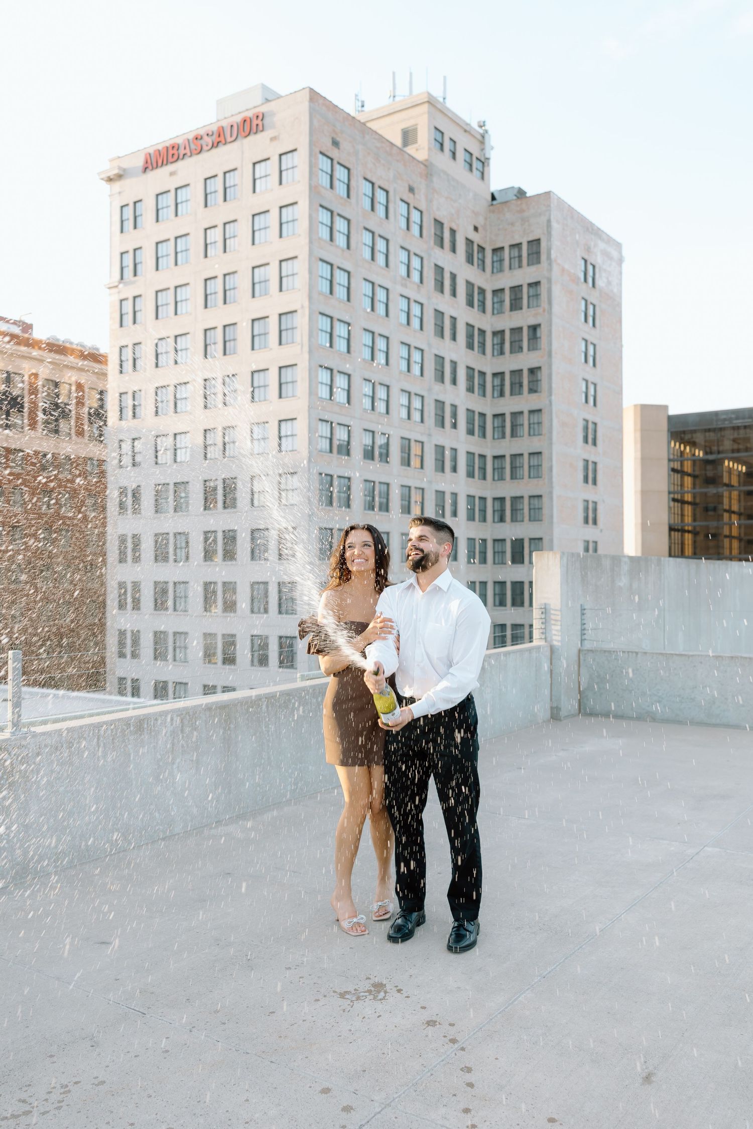 Couple popping champagne bottle on rooftop with city skyline backdrop, celebrating their engagement.