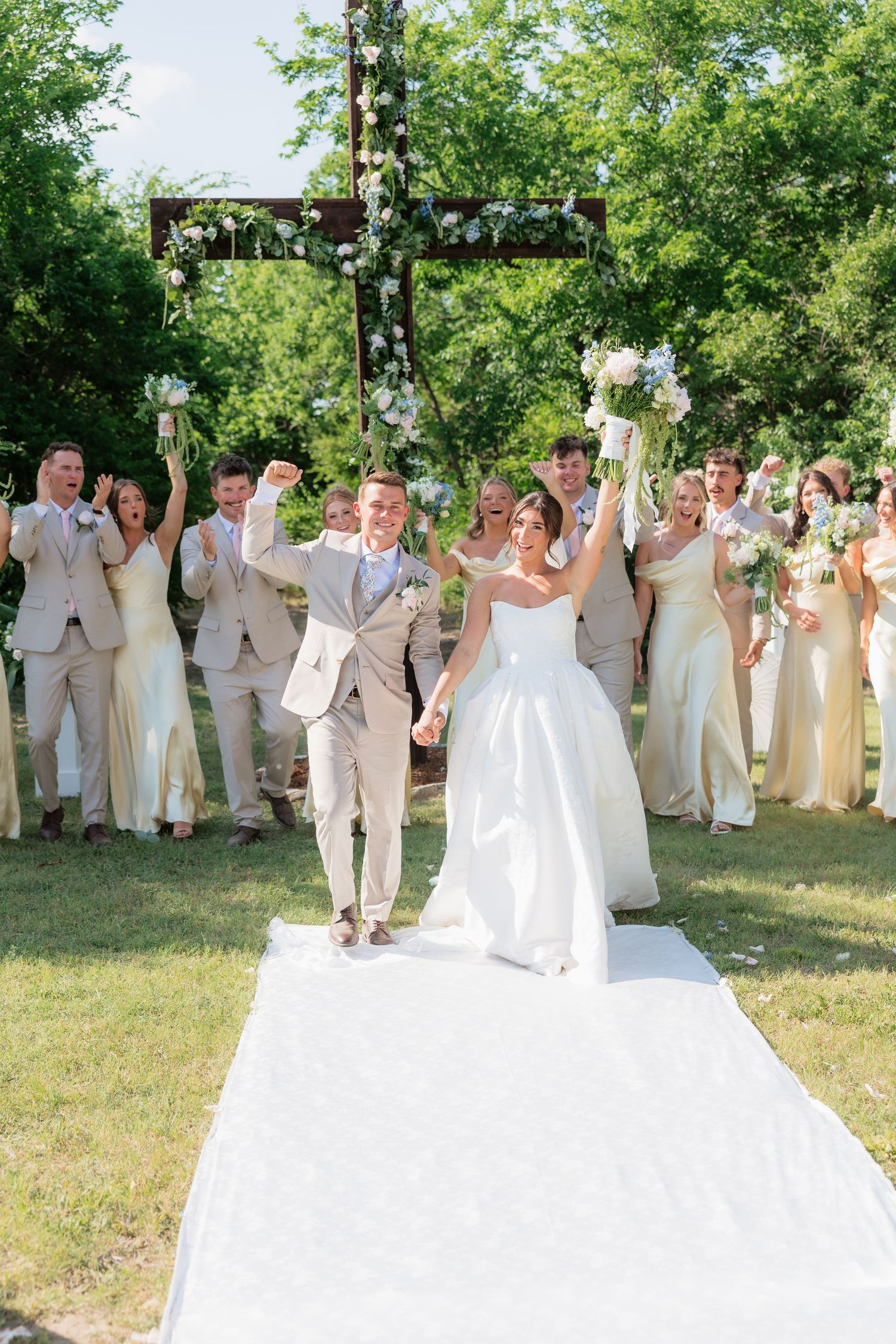 Joyful bride and groom walk back down aisle as wedding party celebrates behind them outdoors.