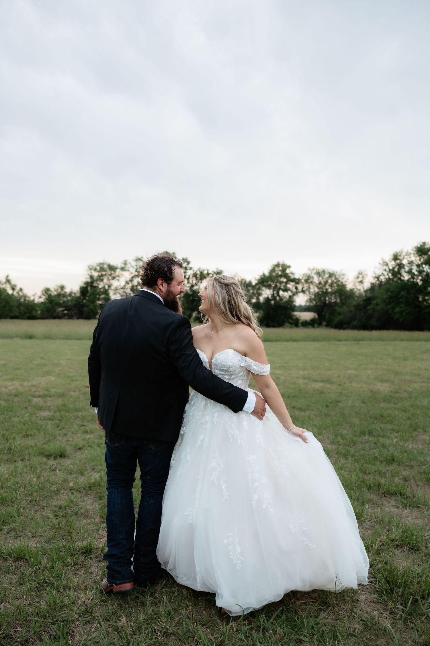 Bride and groom walk together through open green field at dusk after outdoor wedding ceremony.
