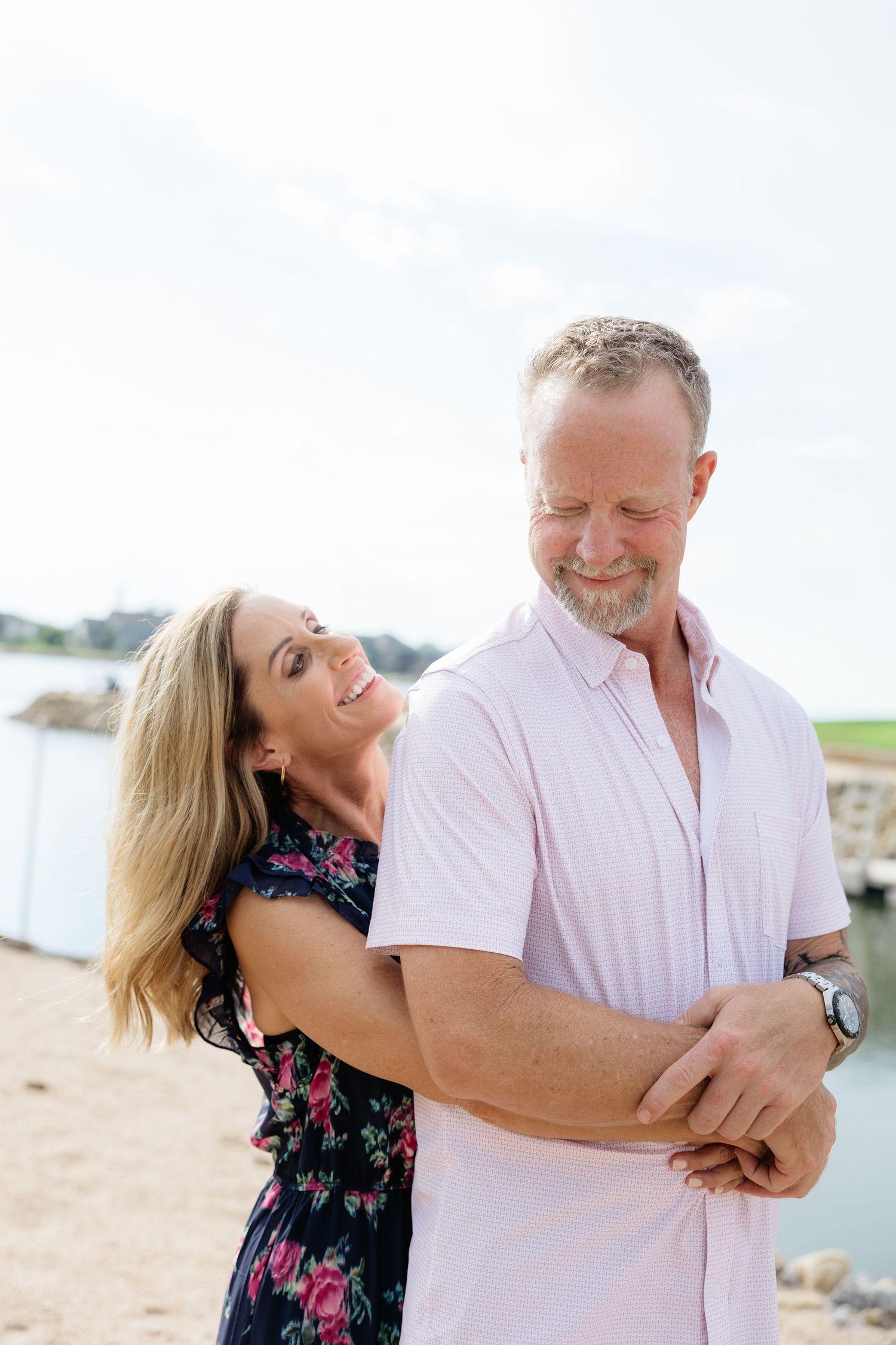 Woman embracing man from behind as they smile together near a waterfront beach.