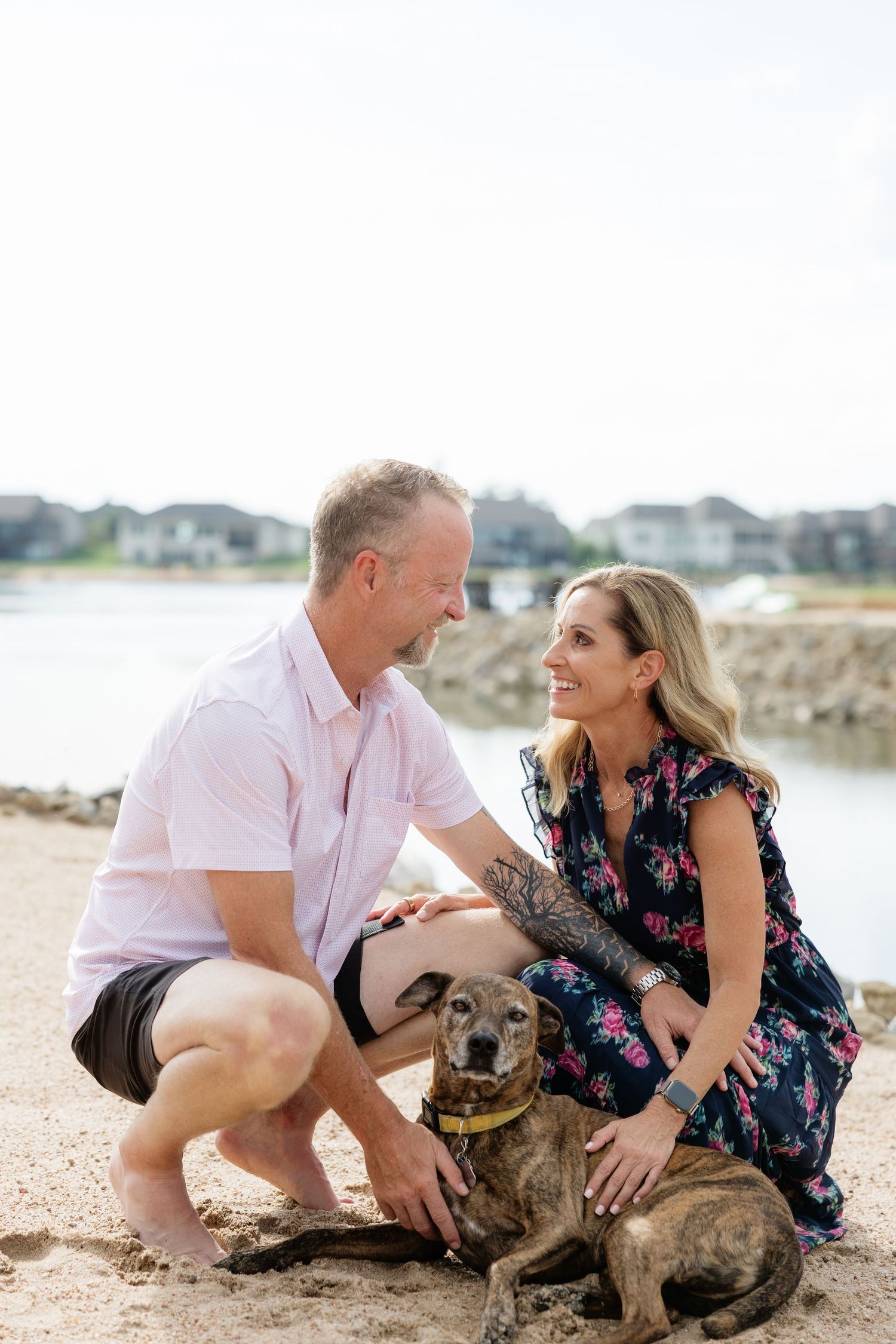 Couple smiling at each other while petting their brindle dog on a sandy beach.