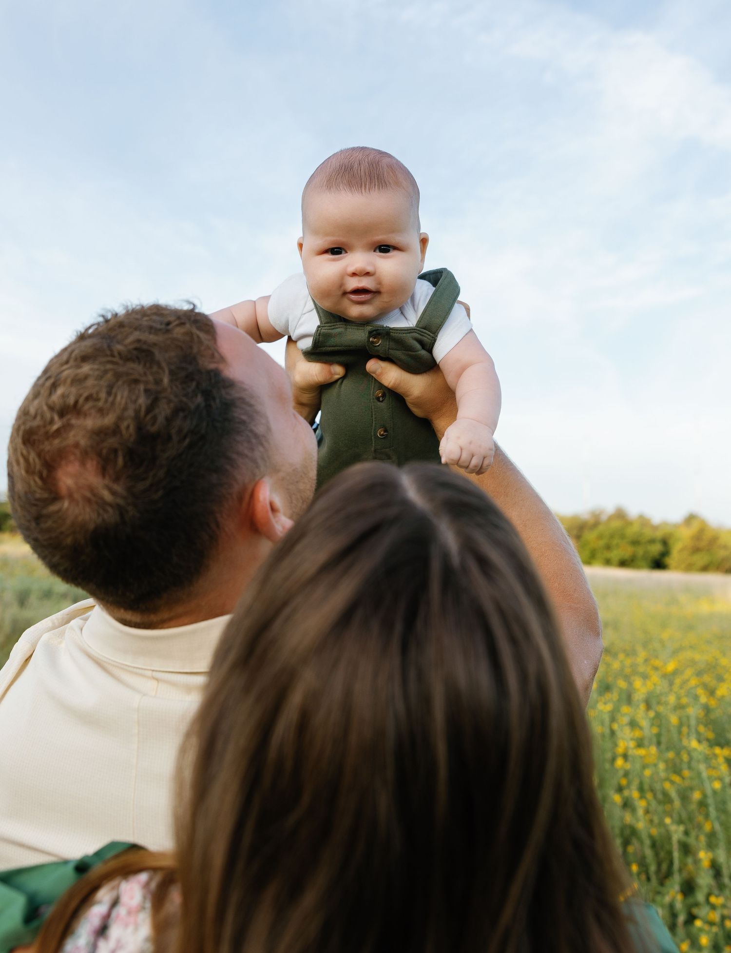 Parents joyfully lifting their smiling baby in a sunny wildflower field.
