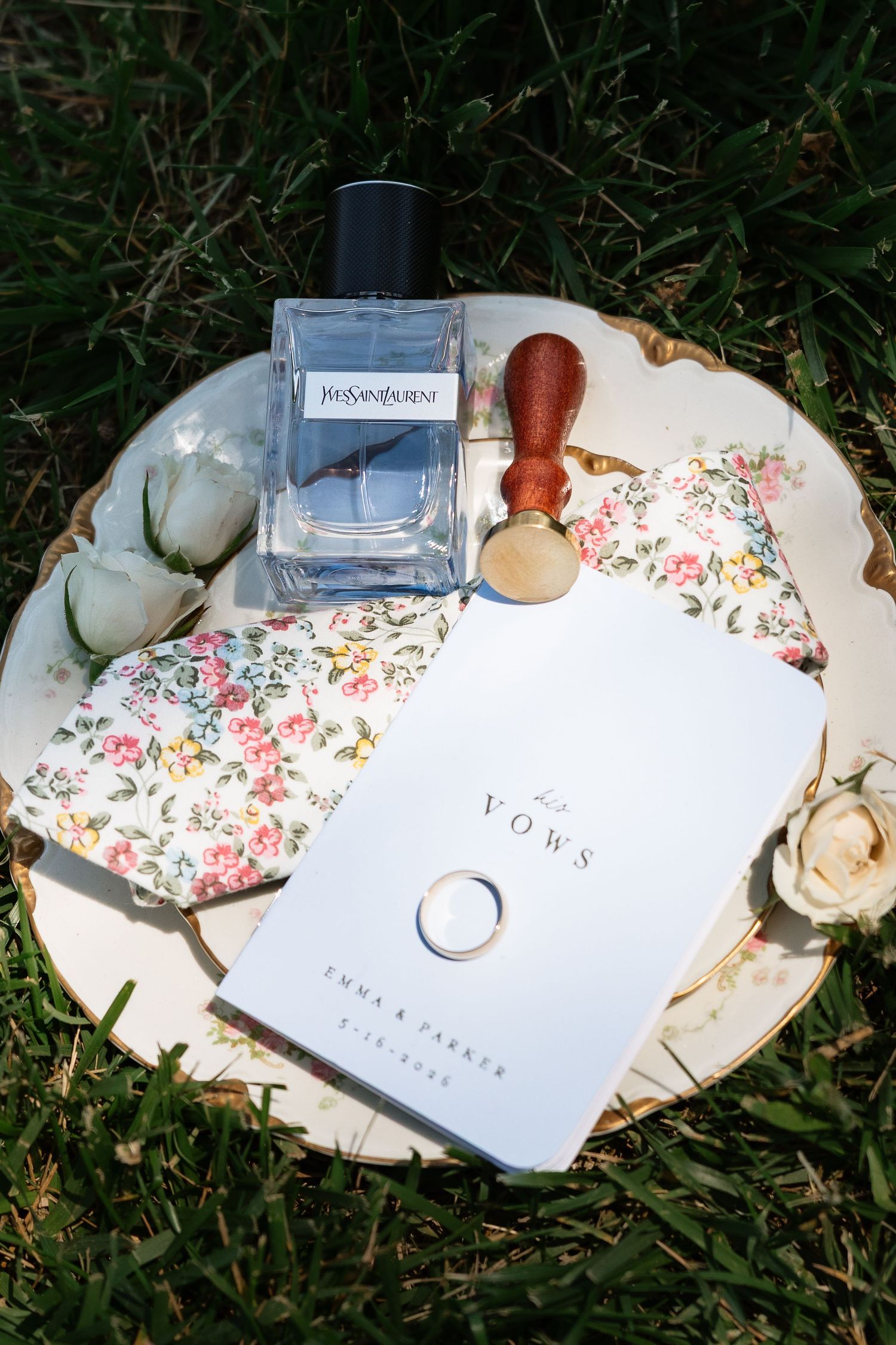 Wedding detail flat lay with YSL cologne, vow books, ring, wax seal, and floral pocket square.