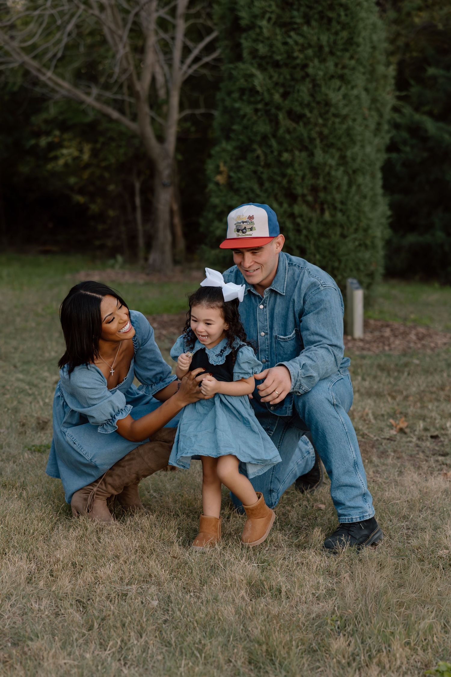 Family in matching denim outfits laughing together on grass with evergreen trees background.