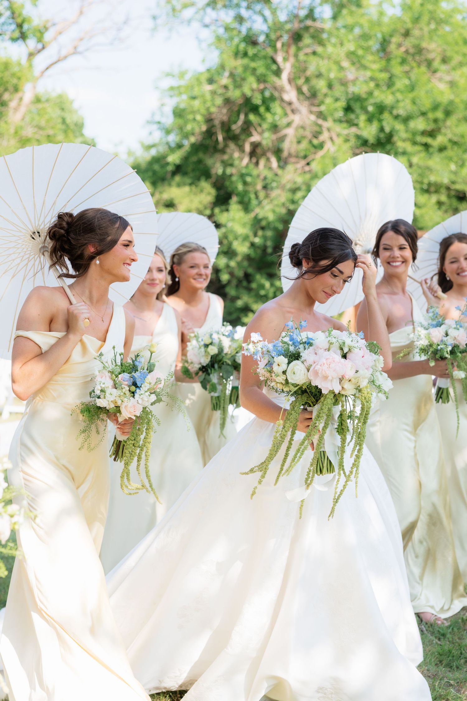 Bride and bridesmaids in ivory and gold gowns walk together holding pastel floral bouquets under white parasols.
