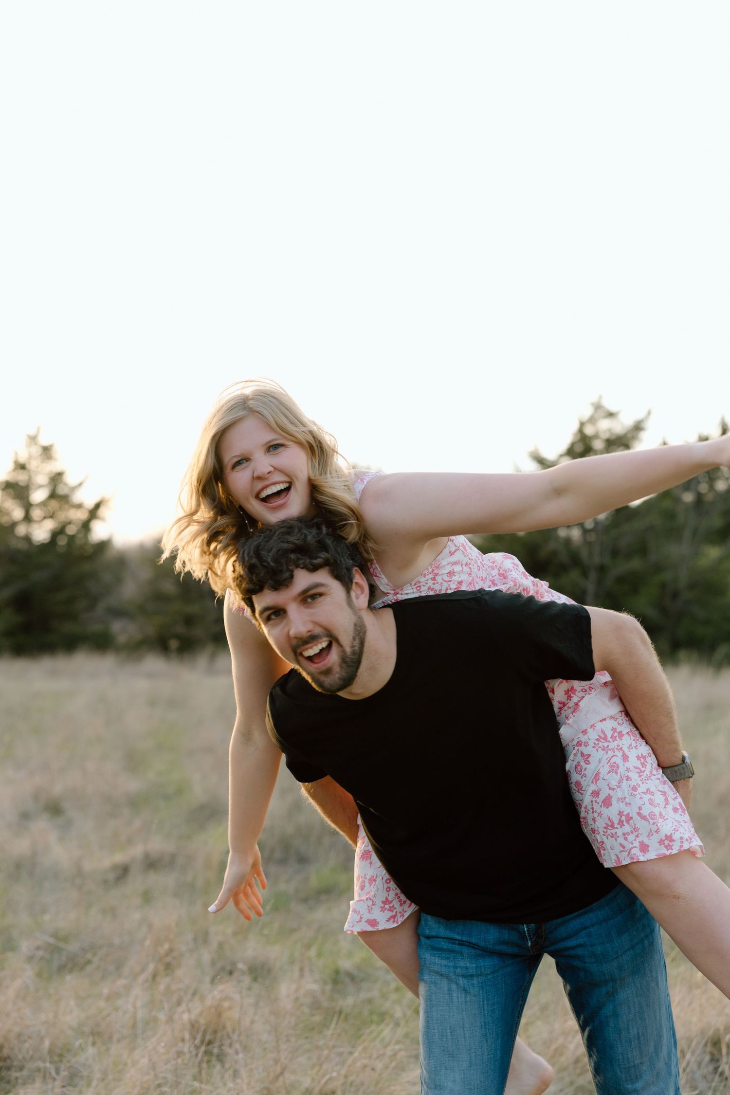 Joyful couple playing outdoors, man giving woman a piggyback ride in a golden field at sunset.