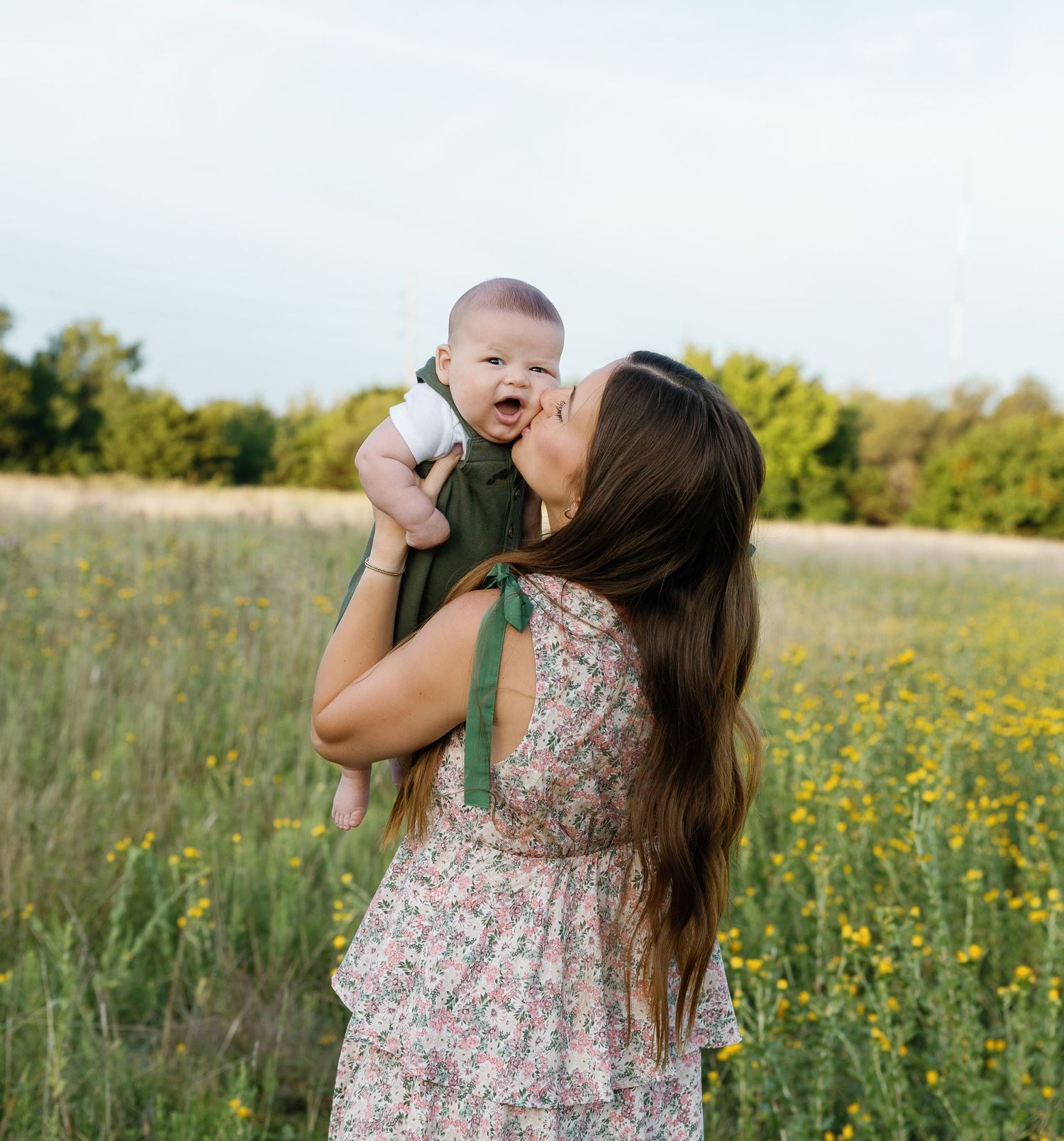 Mother kissing her baby while holding them up in a green meadow.