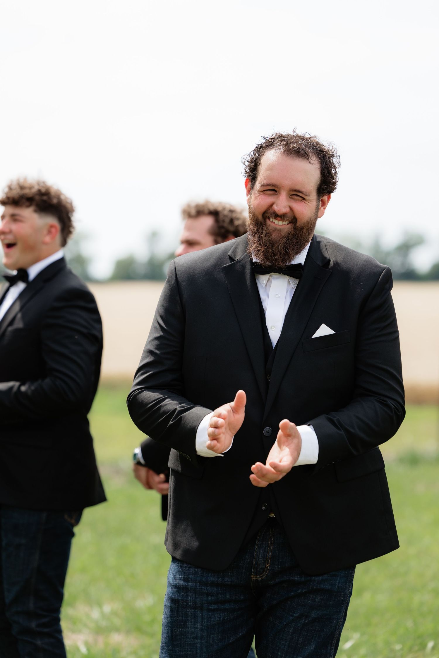 Bearded groom in black tuxedo and bow tie smiles and claps joyfully outdoors with groomsmen nearby.