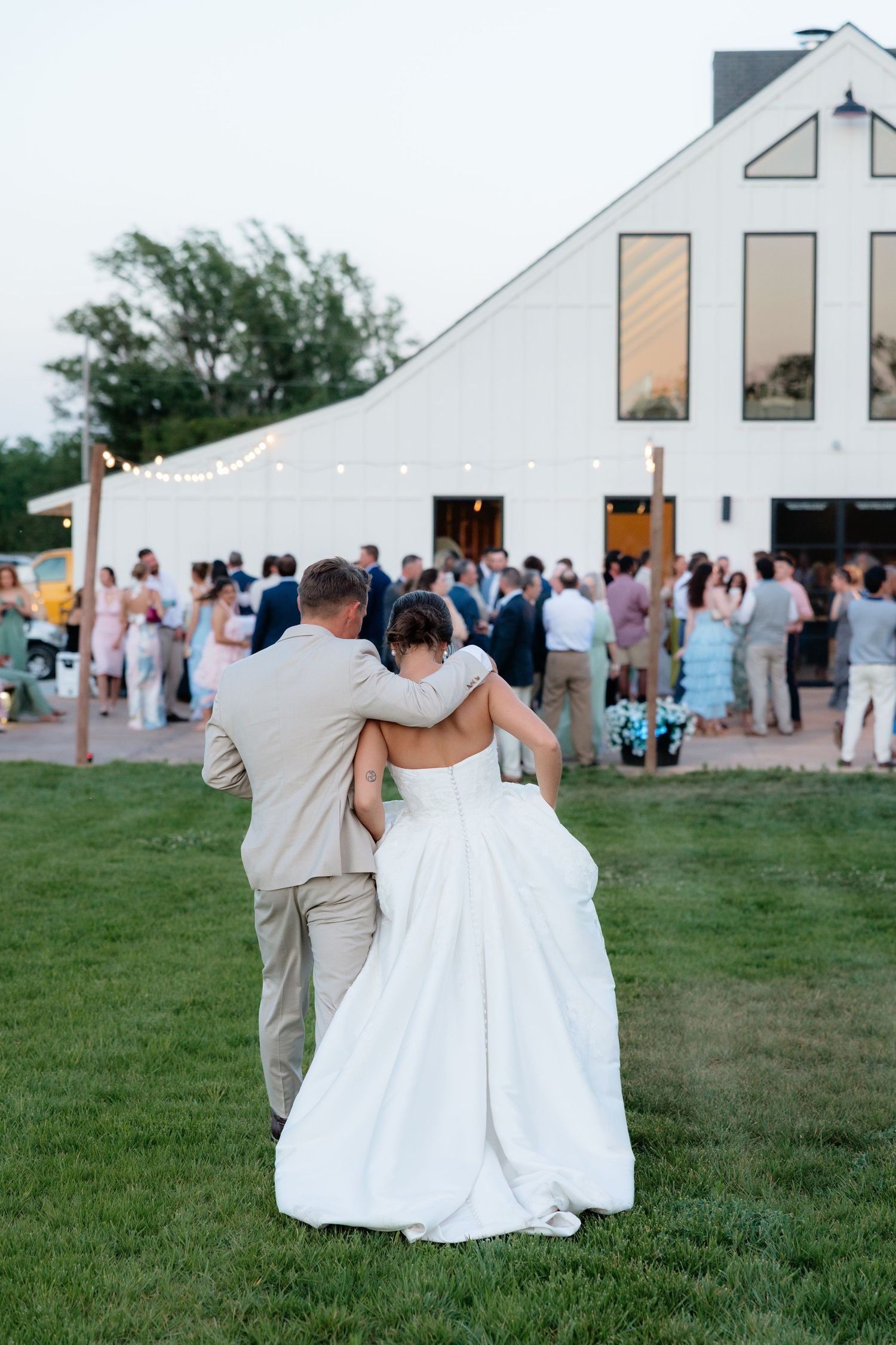 Bride and groom walking arm in arm toward white barn reception venue filled with celebrating wedding guests.