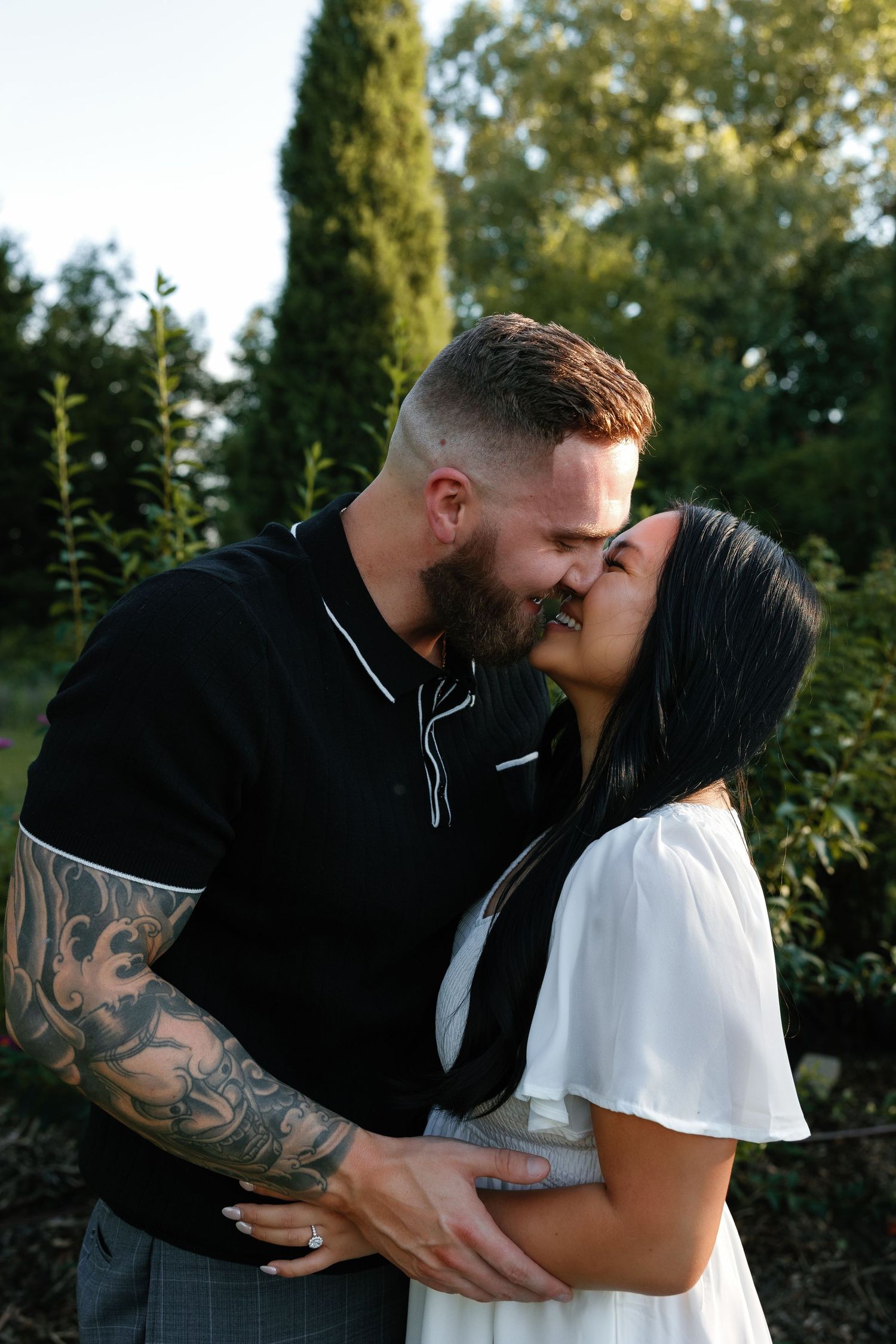 Tattooed couple sharing a kiss outdoors at dusk surrounded by lush green trees and cypress hedges.