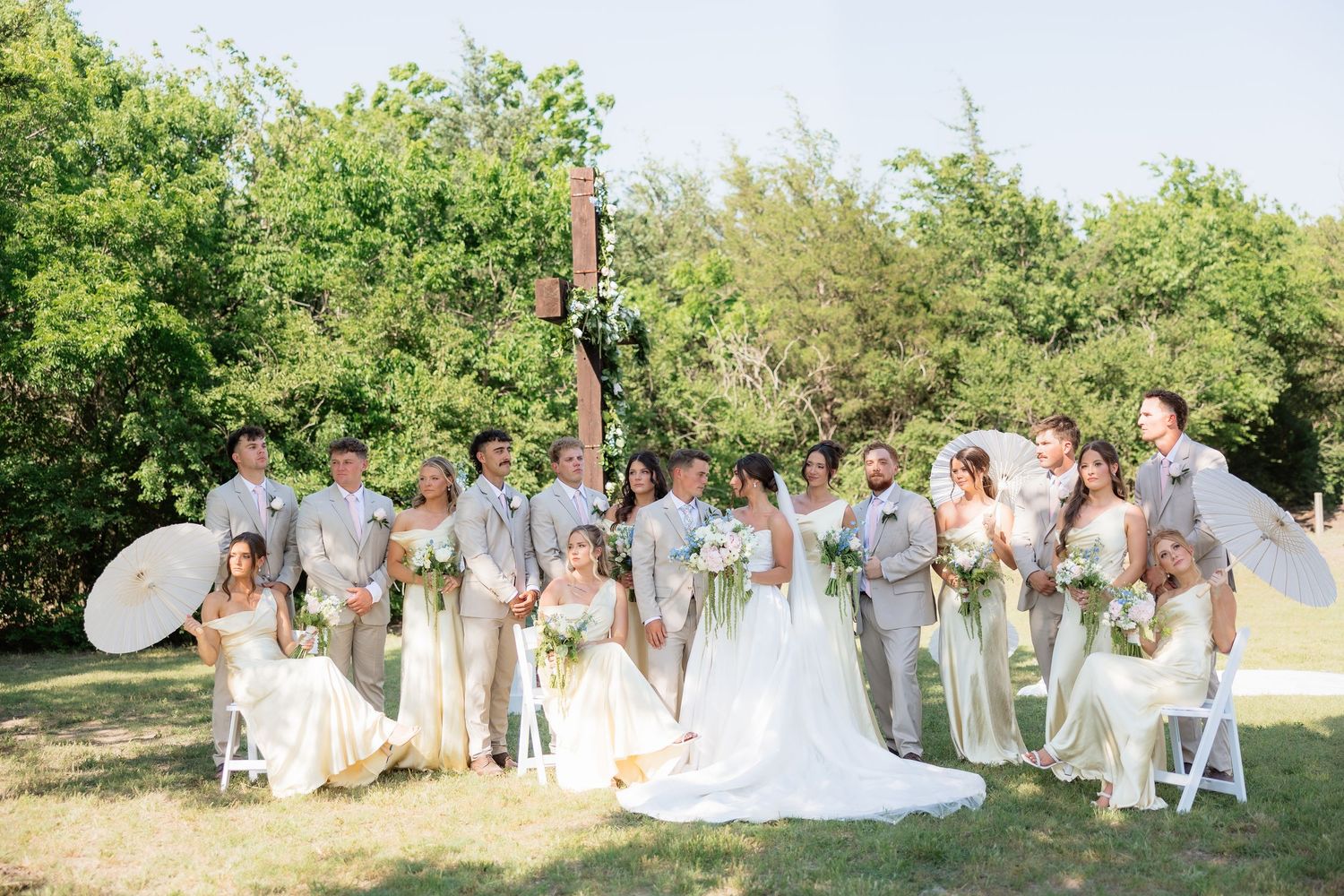 Wedding party poses together outdoors with white parasols near floral cross ceremony backdrop.
