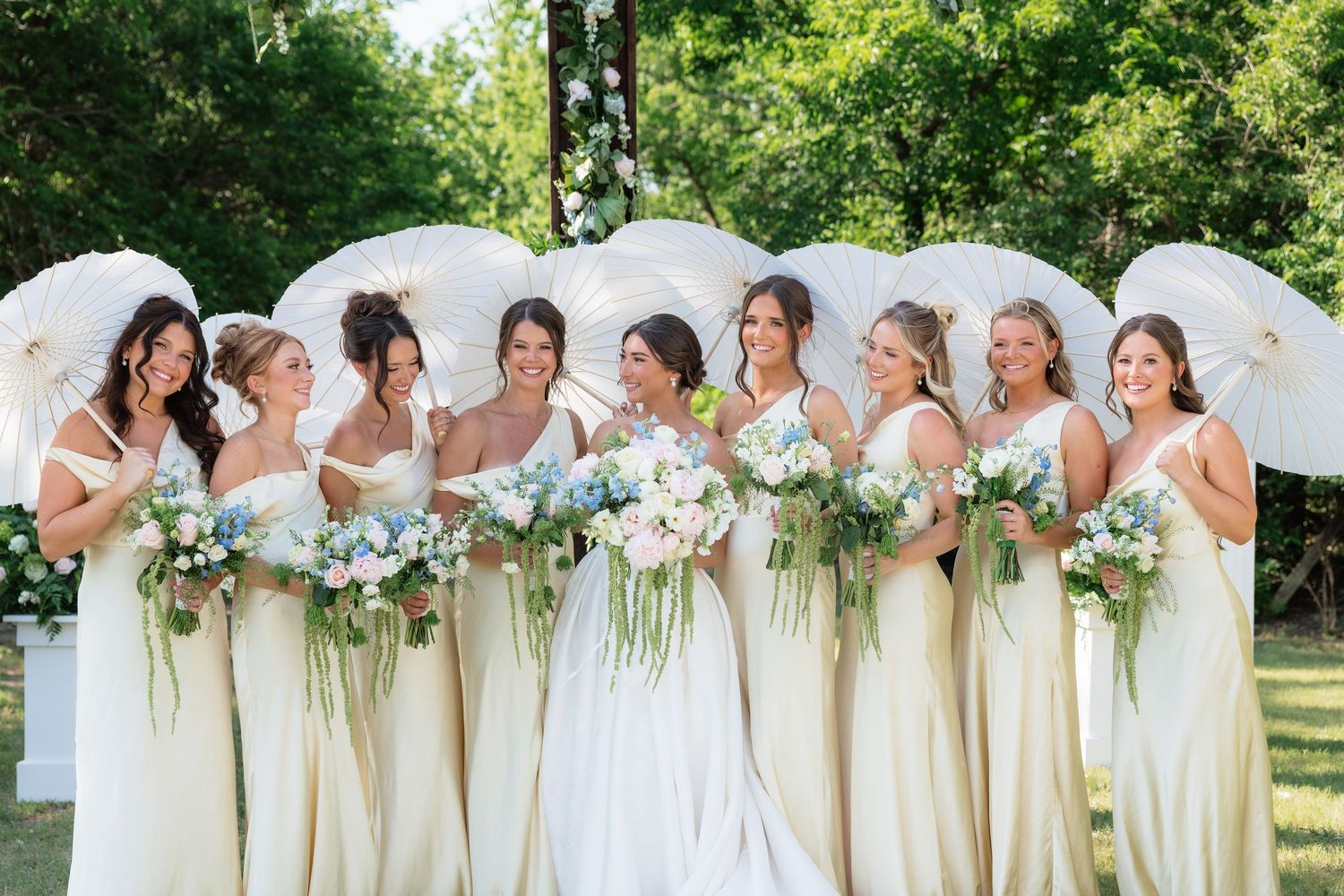 Bride poses with eight bridesmaids in champagne gowns holding floral bouquets and white parasols.