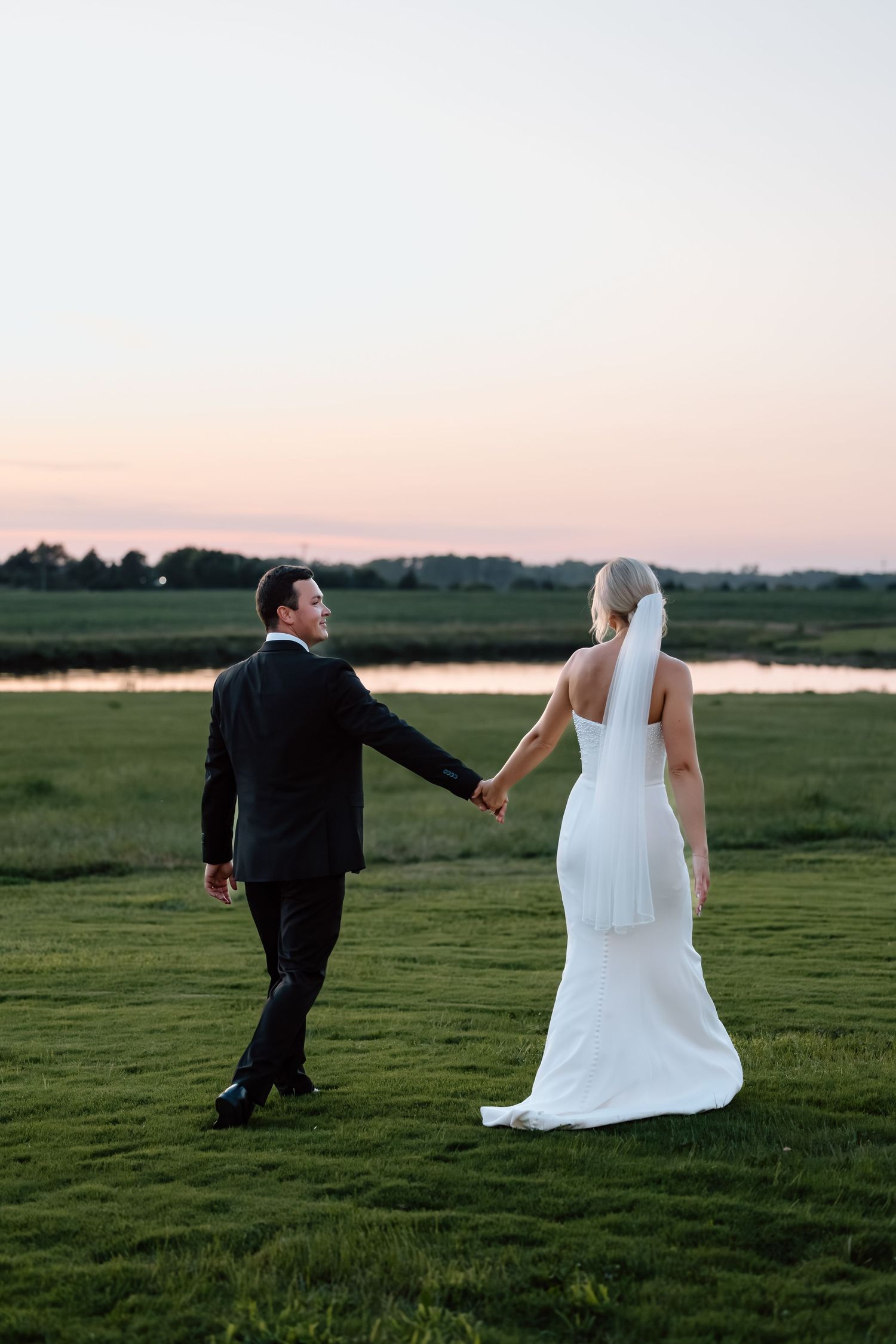 Bride in white halter gown and groom in black tuxedo walking hand in hand across a green field at sunset.