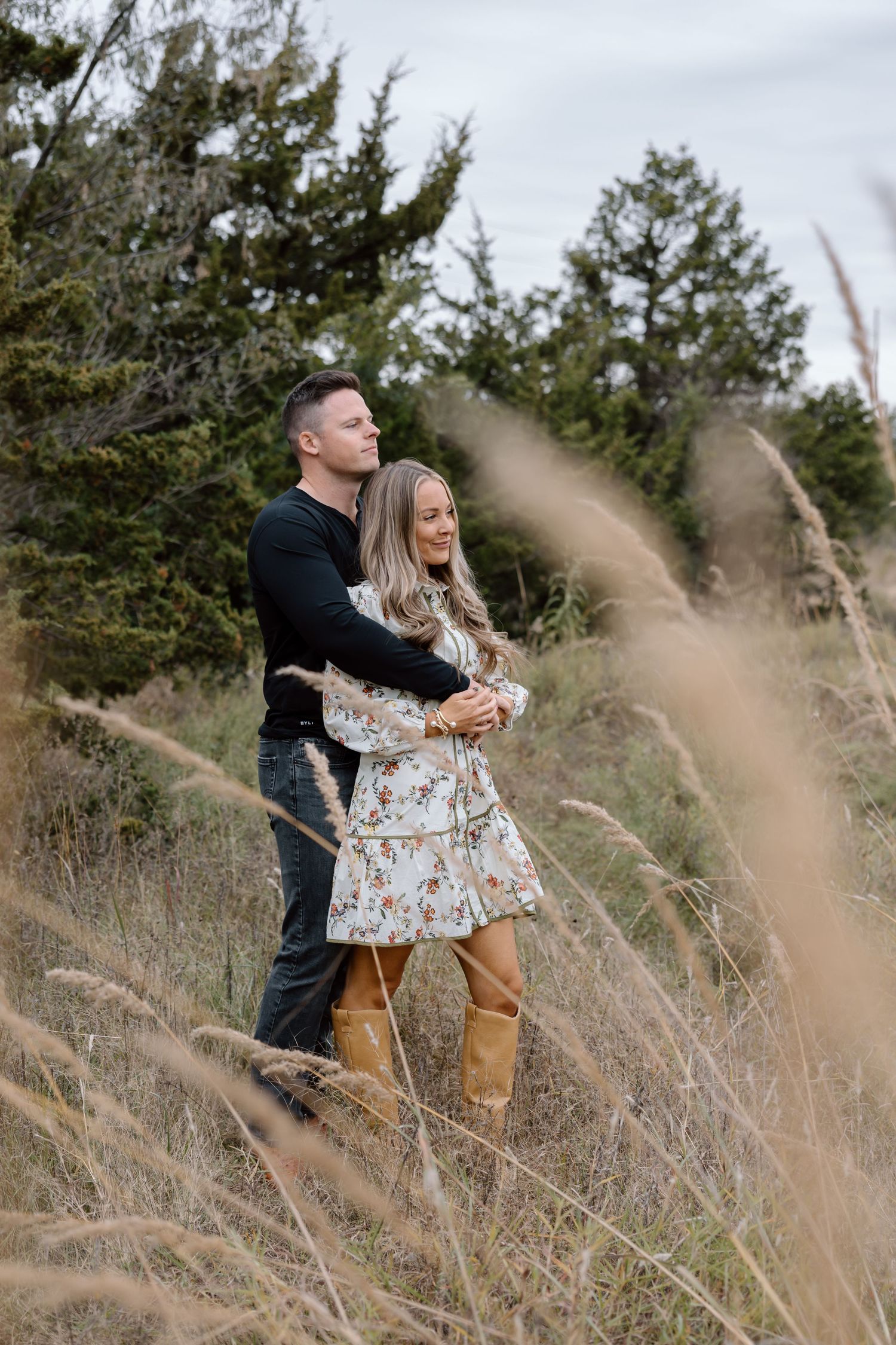 Couple embracing in a golden meadow with evergreen trees in the background.