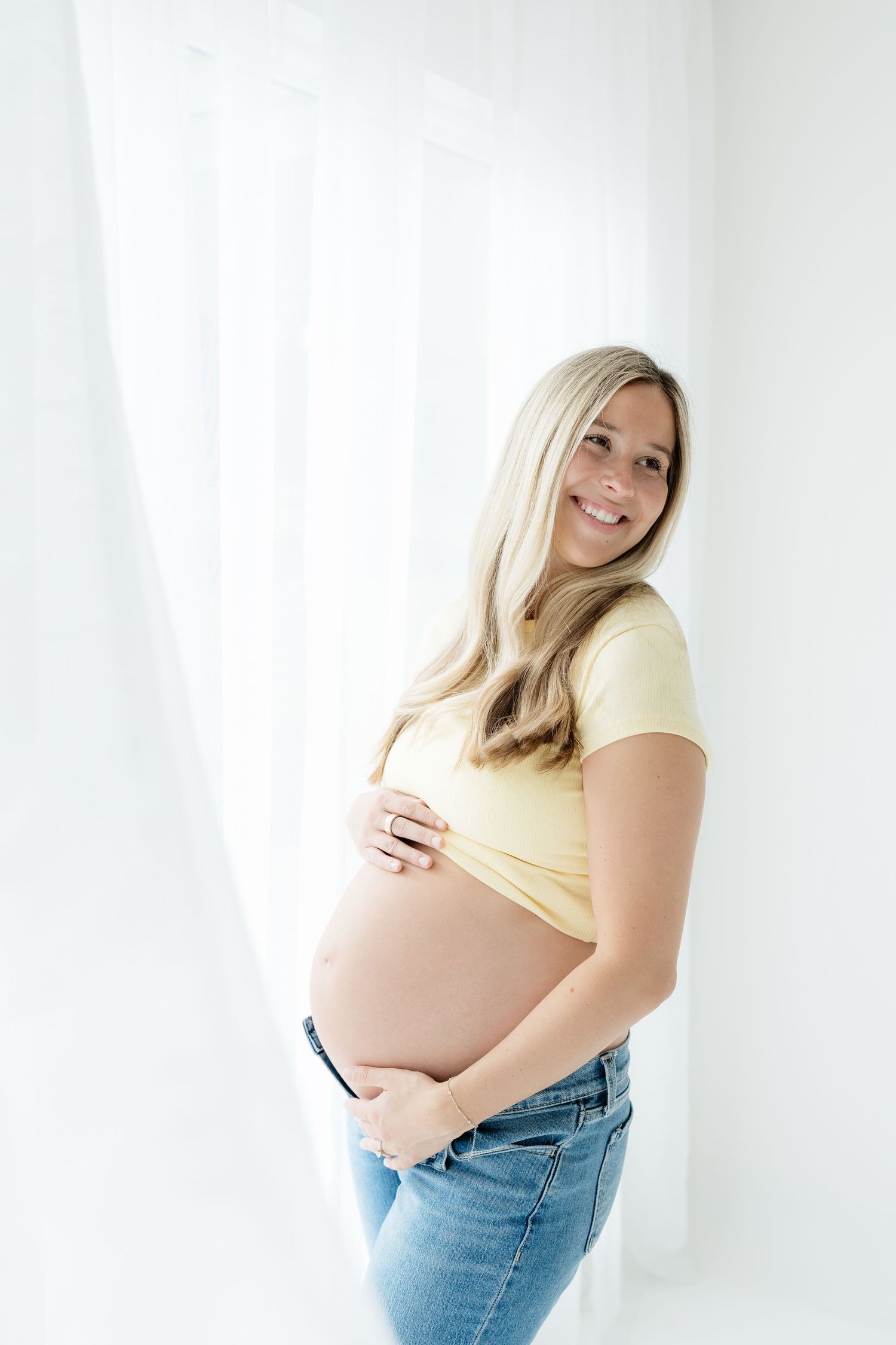 Smiling pregnant woman in yellow crop top and jeans cradling her baby bump in front of white curtains.
