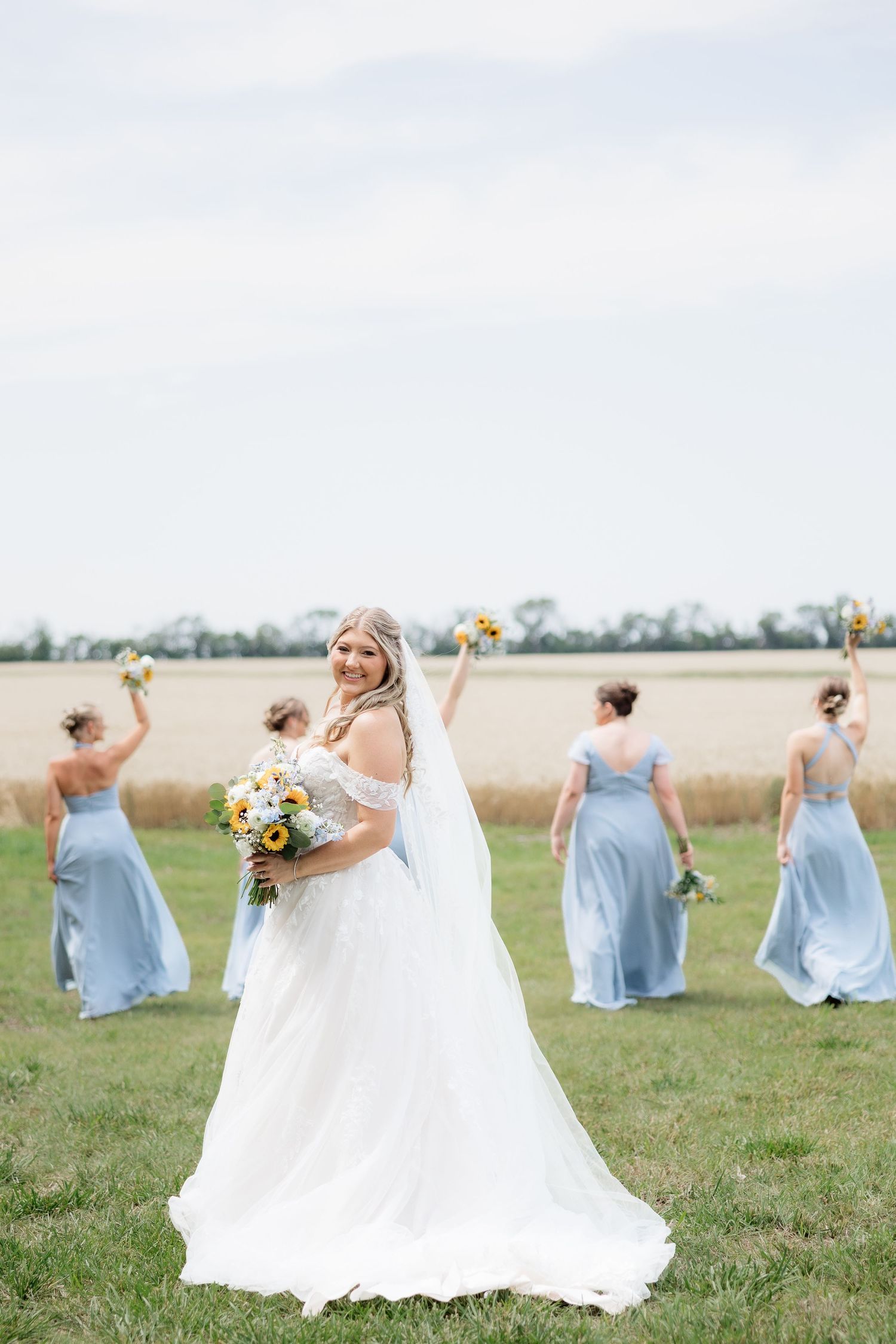 Bride holds sunflower bouquet as bridesmaids in blue dresses celebrate with raised arms in an open field.