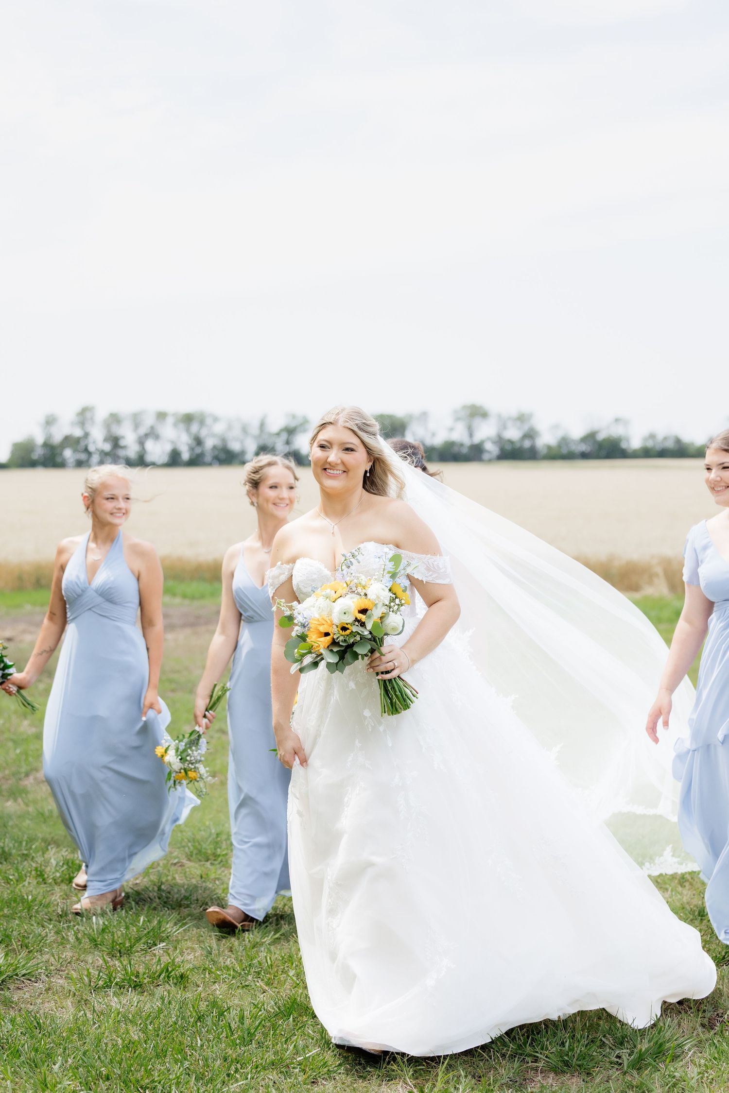 Bride in white lace gown walks with bridesmaids in light blue dresses through a green field on wedding day.