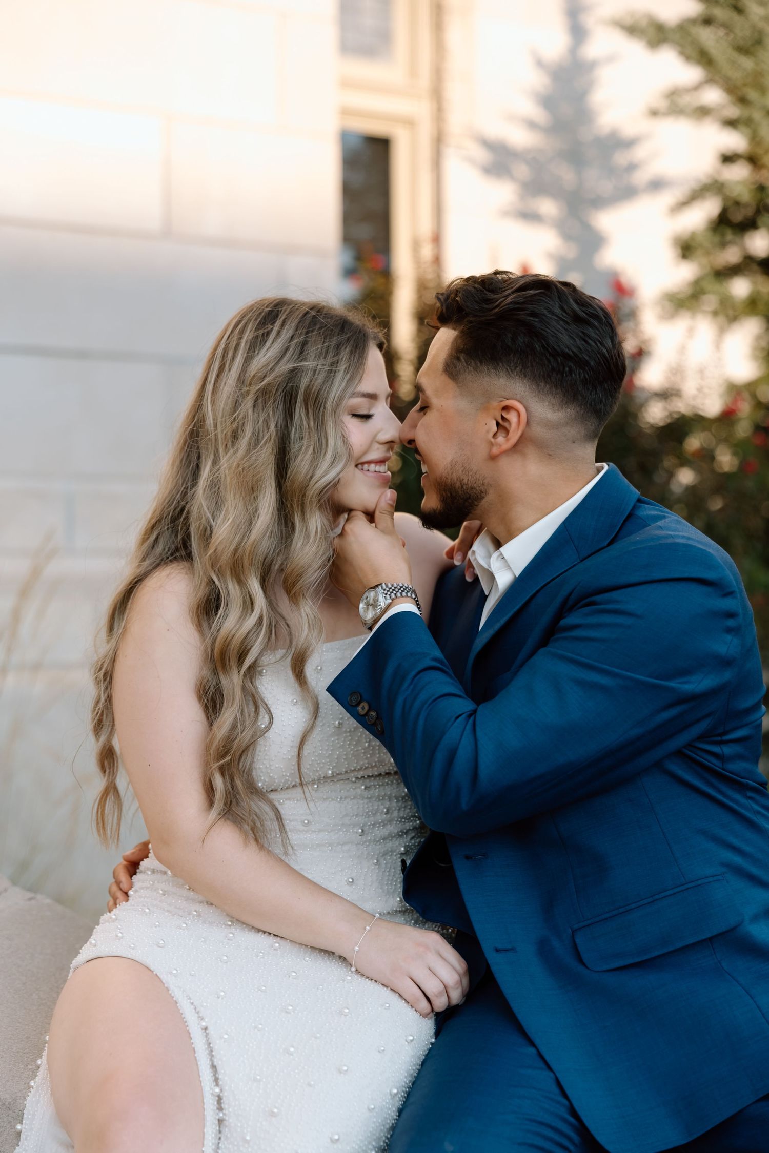 Groom in blue suit tenderly kisses bride in white beaded dress while sitting together outdoors.