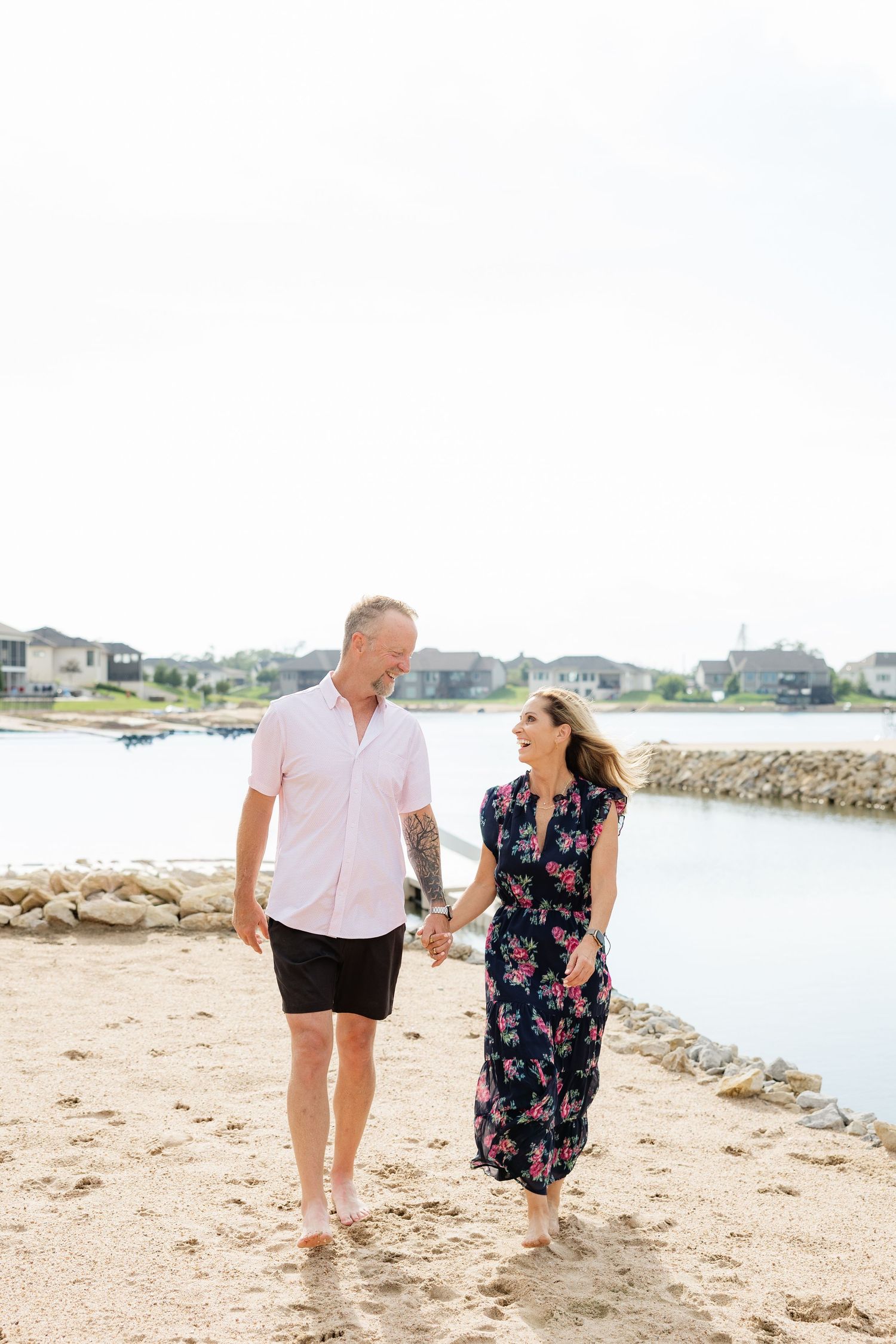 Couple holding hands and smiling while walking along a sunny sandy waterfront beach.