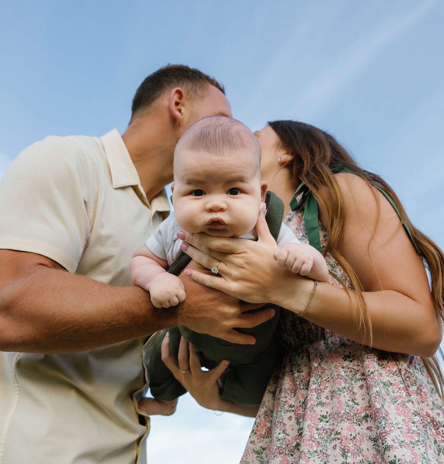 New parents kissing their newborn baby on the cheeks under a bright blue sky.