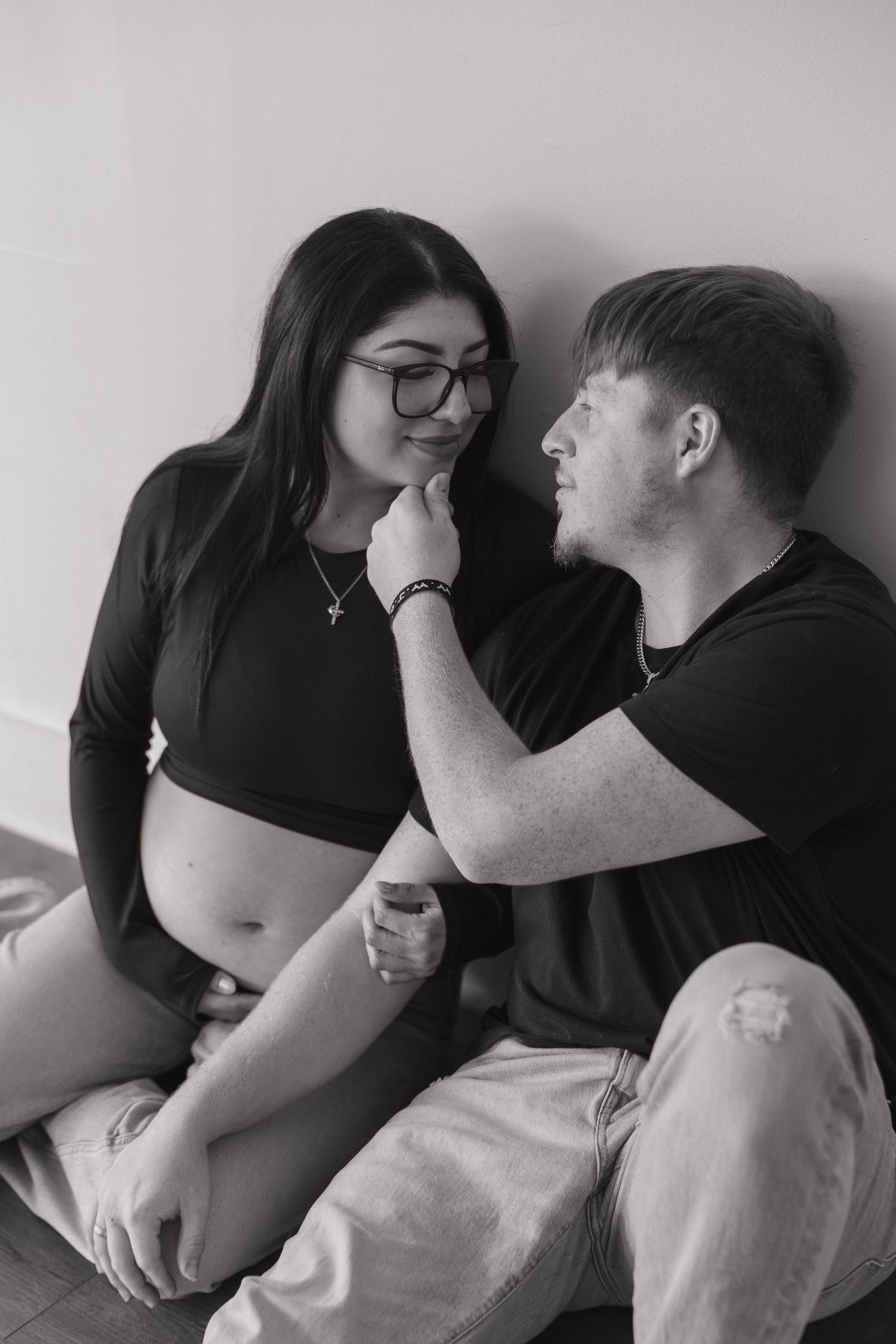 A black and white portrait of a pregnant woman and her partner sharing a tender moment seated against a wall.