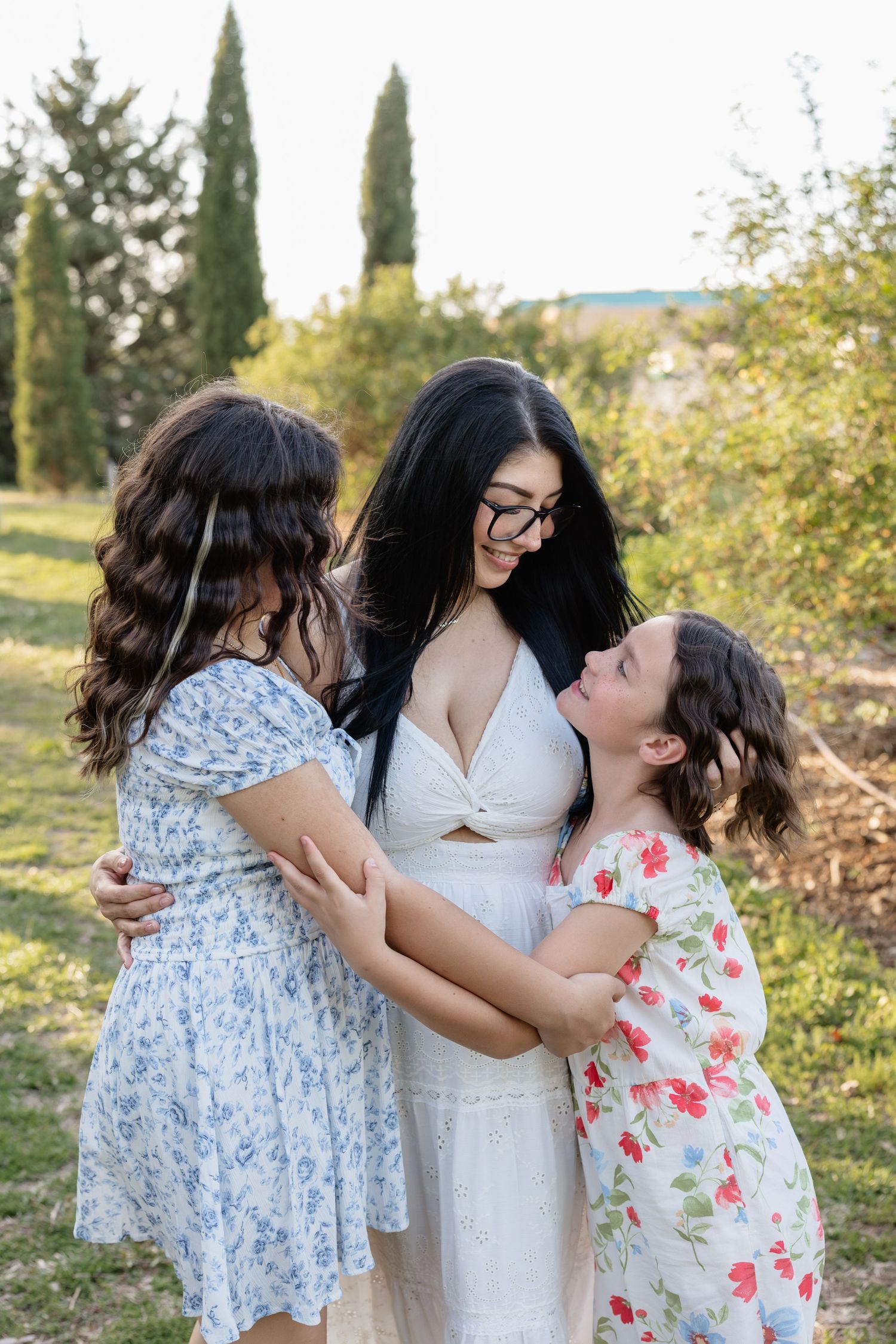 A smiling dark-haired woman in white dress hugs two young girls in floral dresses in a sunny outdoor garden setting.
