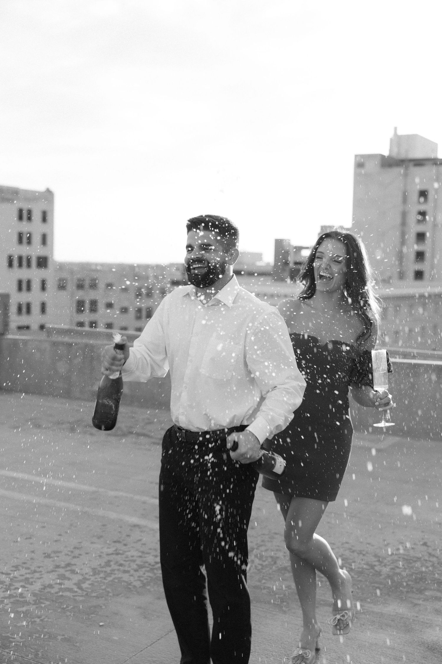 Black and white photo of a joyful couple celebrating with champagne on a rooftop with city buildings in background.