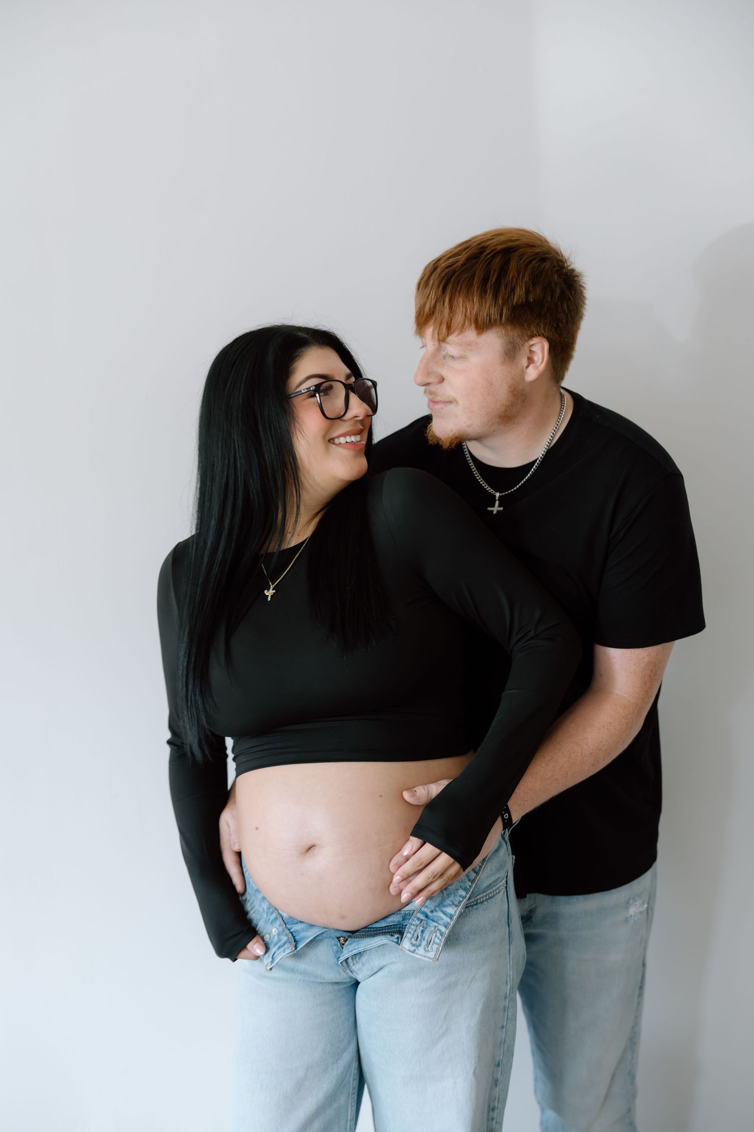 A smiling pregnant woman with glasses and her red-haired partner pose together in matching black outfits.