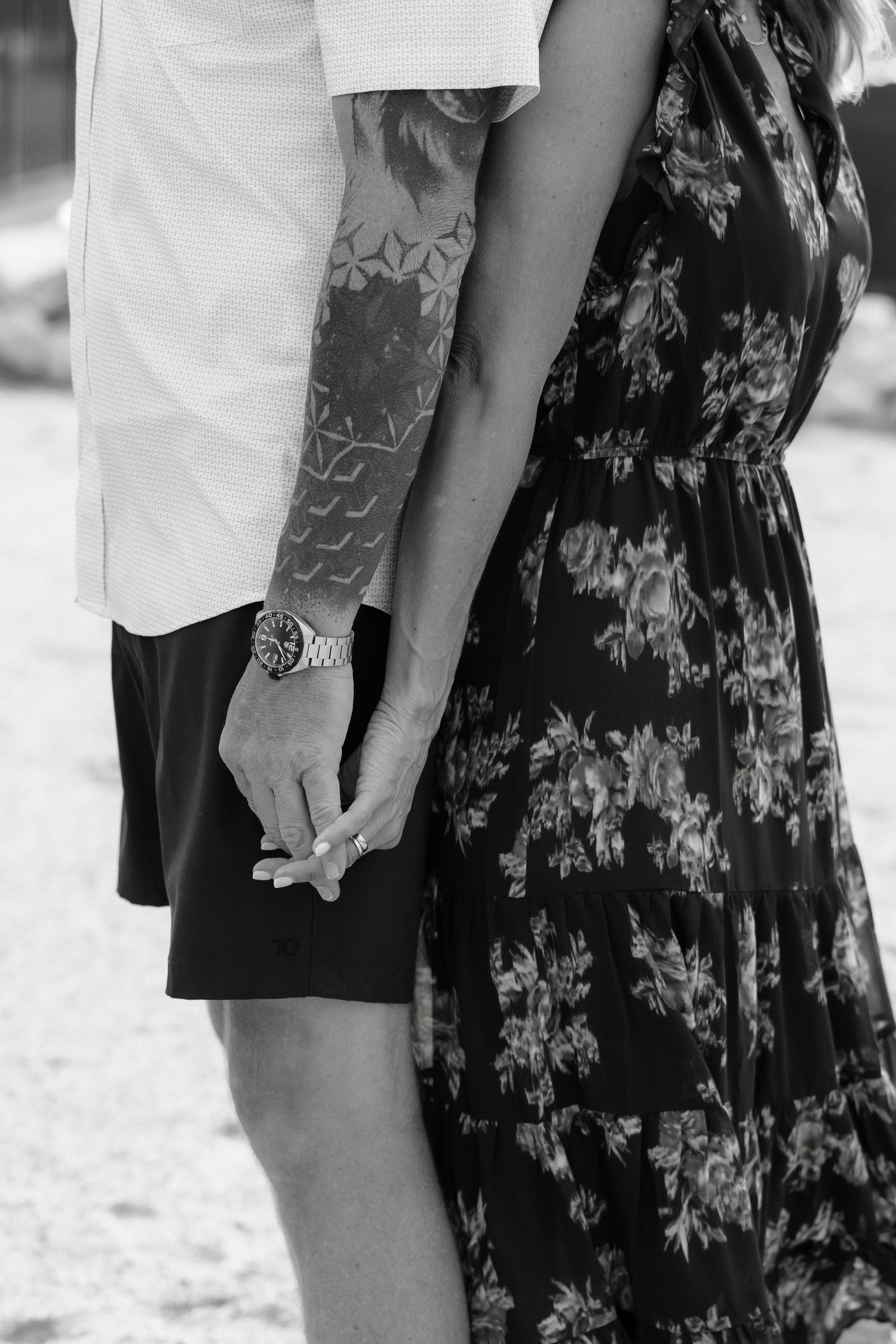 Black and white close-up of tattooed couple holding hands in floral dress outdoors.