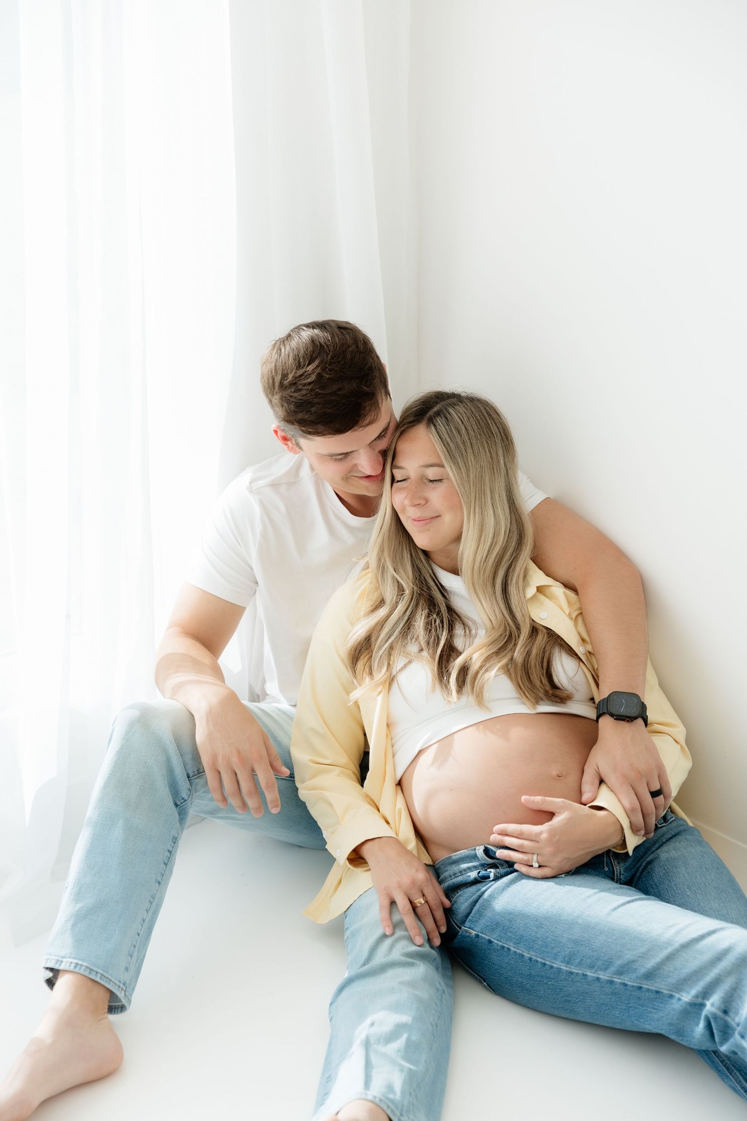 A man in white shirt tenderly holds a pregnant woman from behind as they sit together by a bright window.