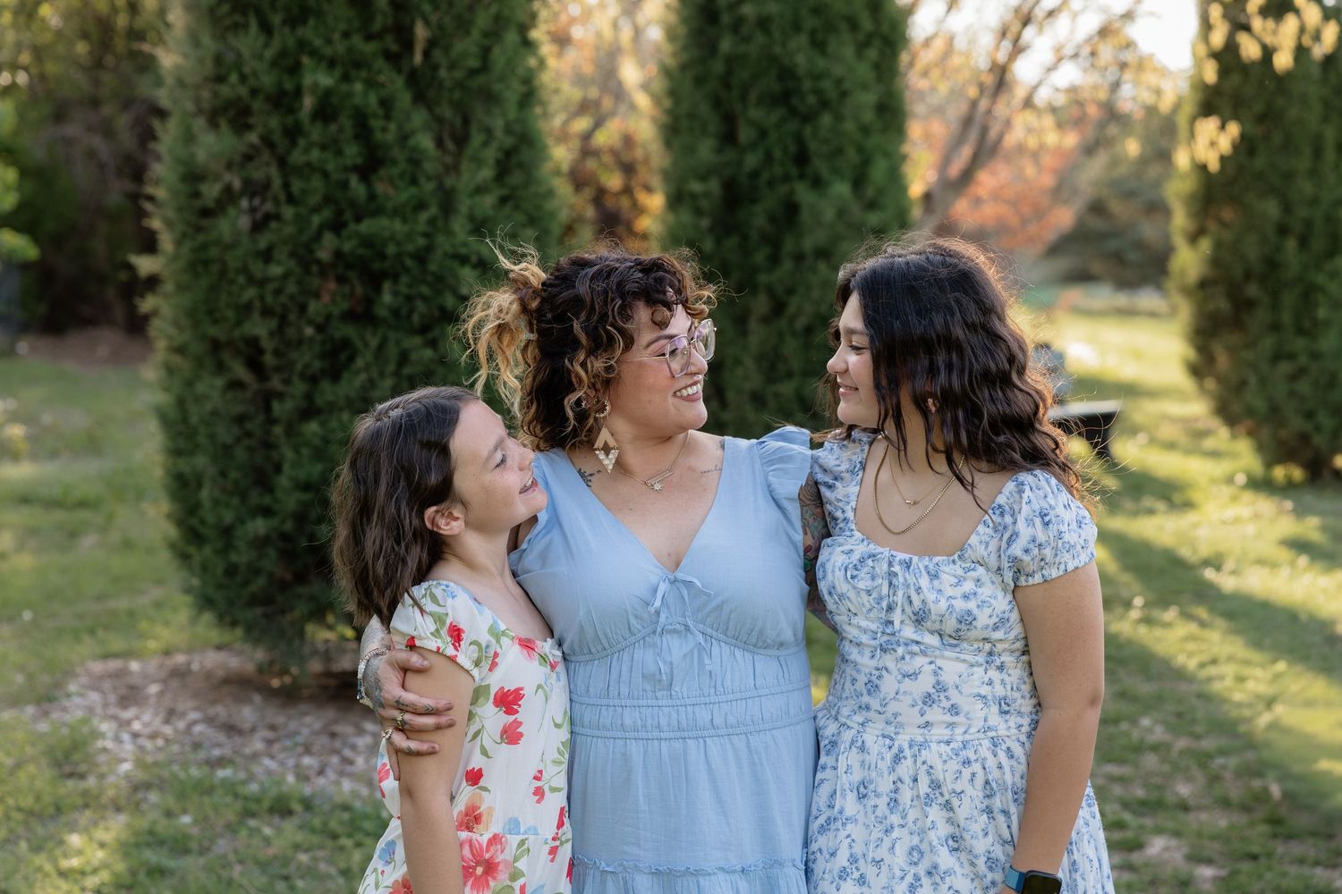 A smiling woman in a blue dress embraces two young girls in floral dresses in a sunny garden setting.