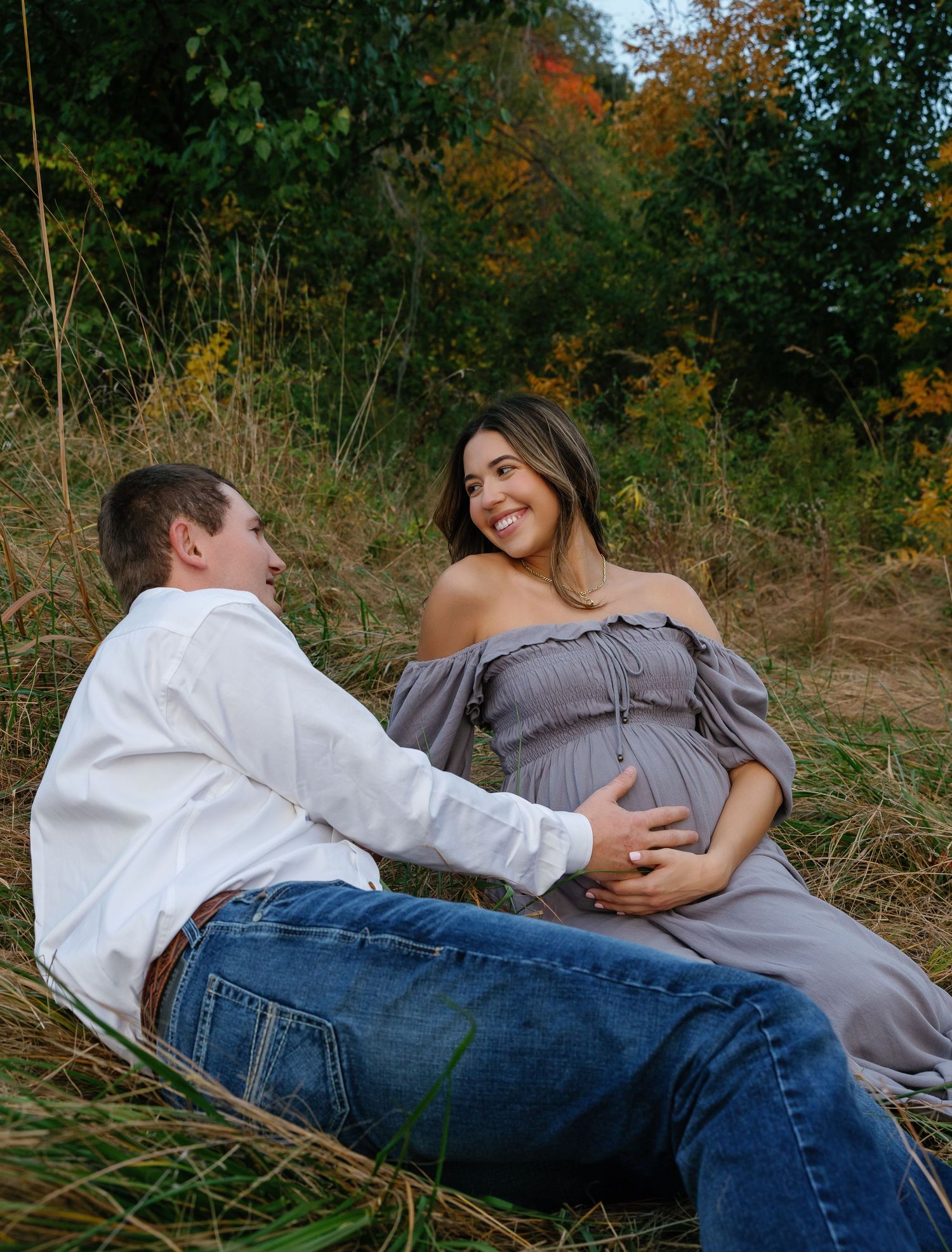 Couple sharing a tender maternity photo session in an autumn field, woman in gray off-shoulder dress cradling baby bump.