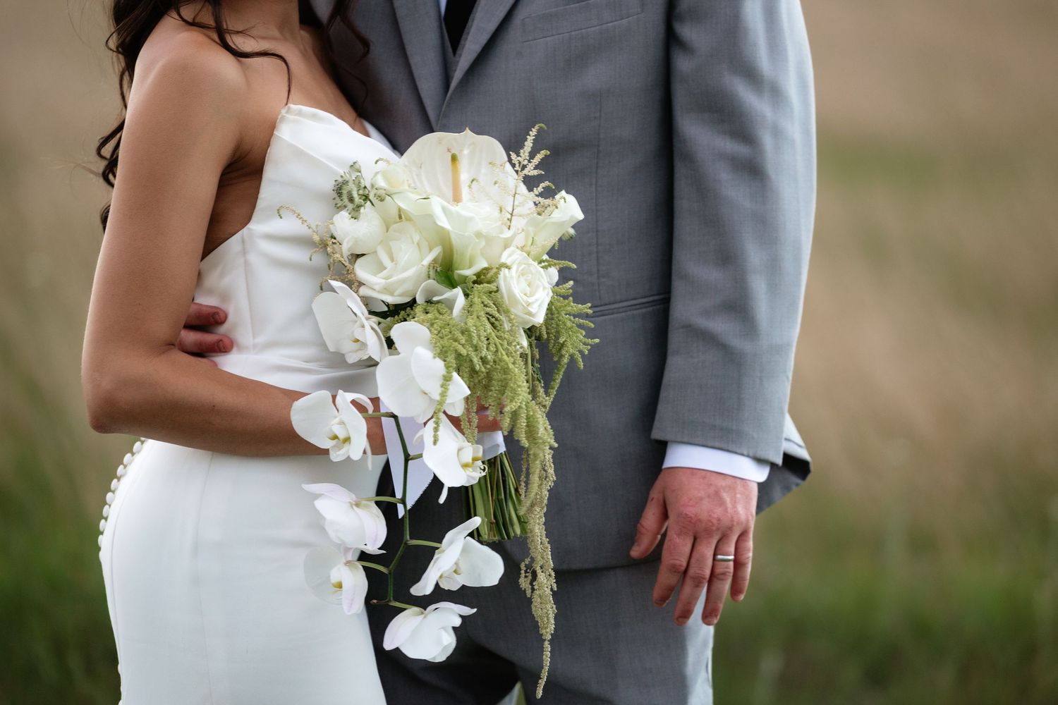 Close-up of bride in white gown holding a cascading white floral bouquet with orchids beside groom in gray suit.