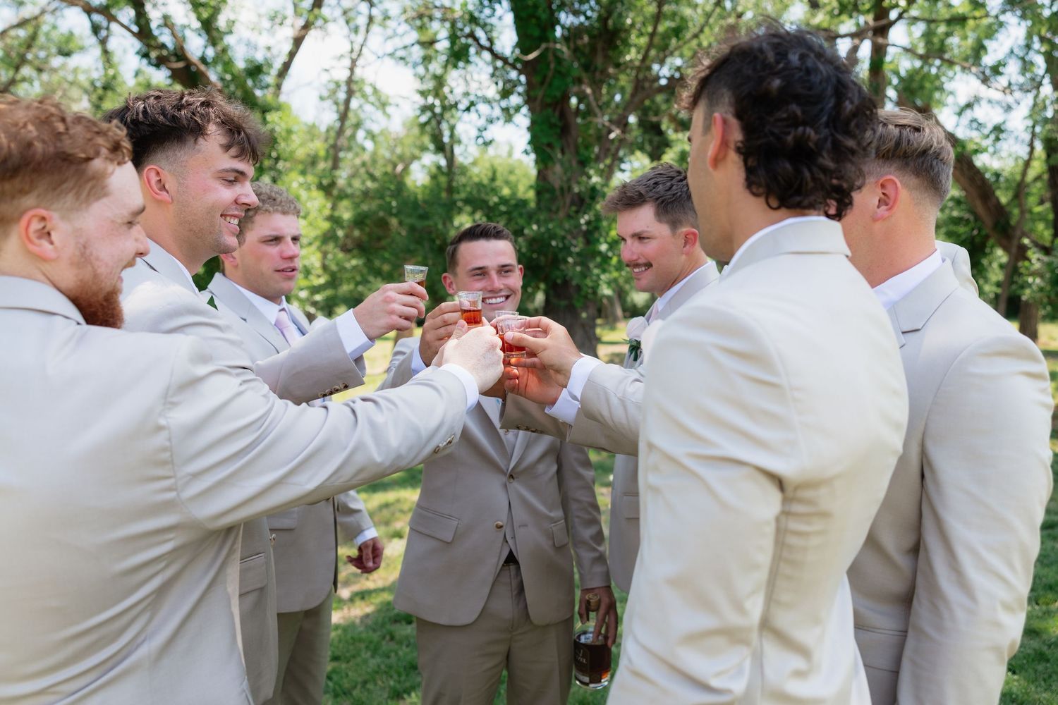 Groomsmen in tan suits raise glasses for a celebratory toast outdoors beneath lush green trees.