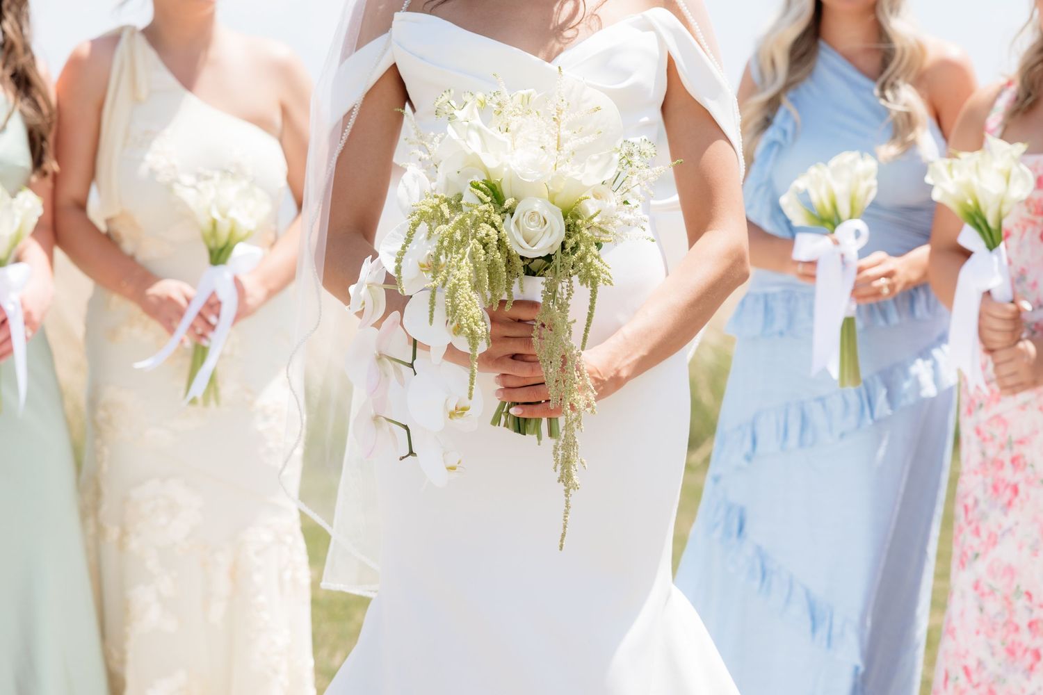 Bride in white off-shoulder gown holding white floral bouquet flanked by bridesmaids in pastel dresses outdoors.