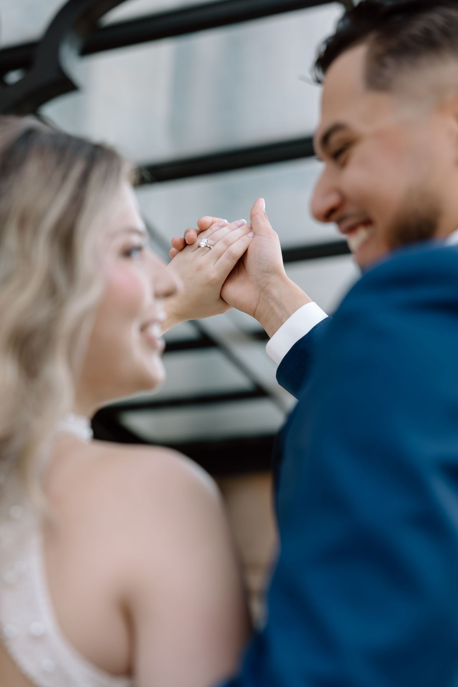 Smiling couple holding hands showing engagement ring, man in blue suit and woman in white dress.