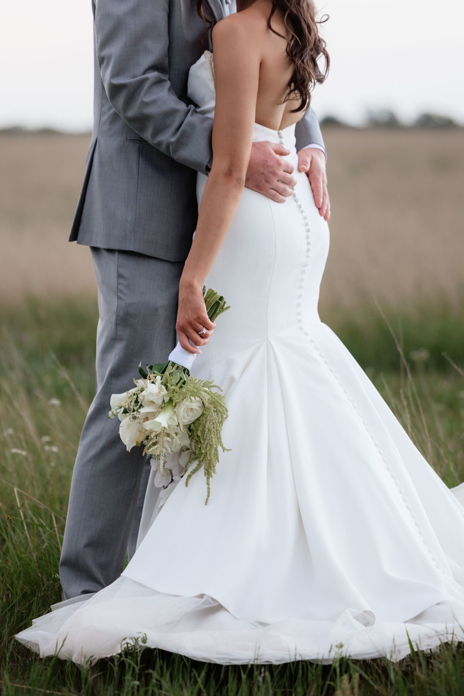 Bride in mermaid wedding gown holding white bouquet embraced by groom in gray suit in field.
