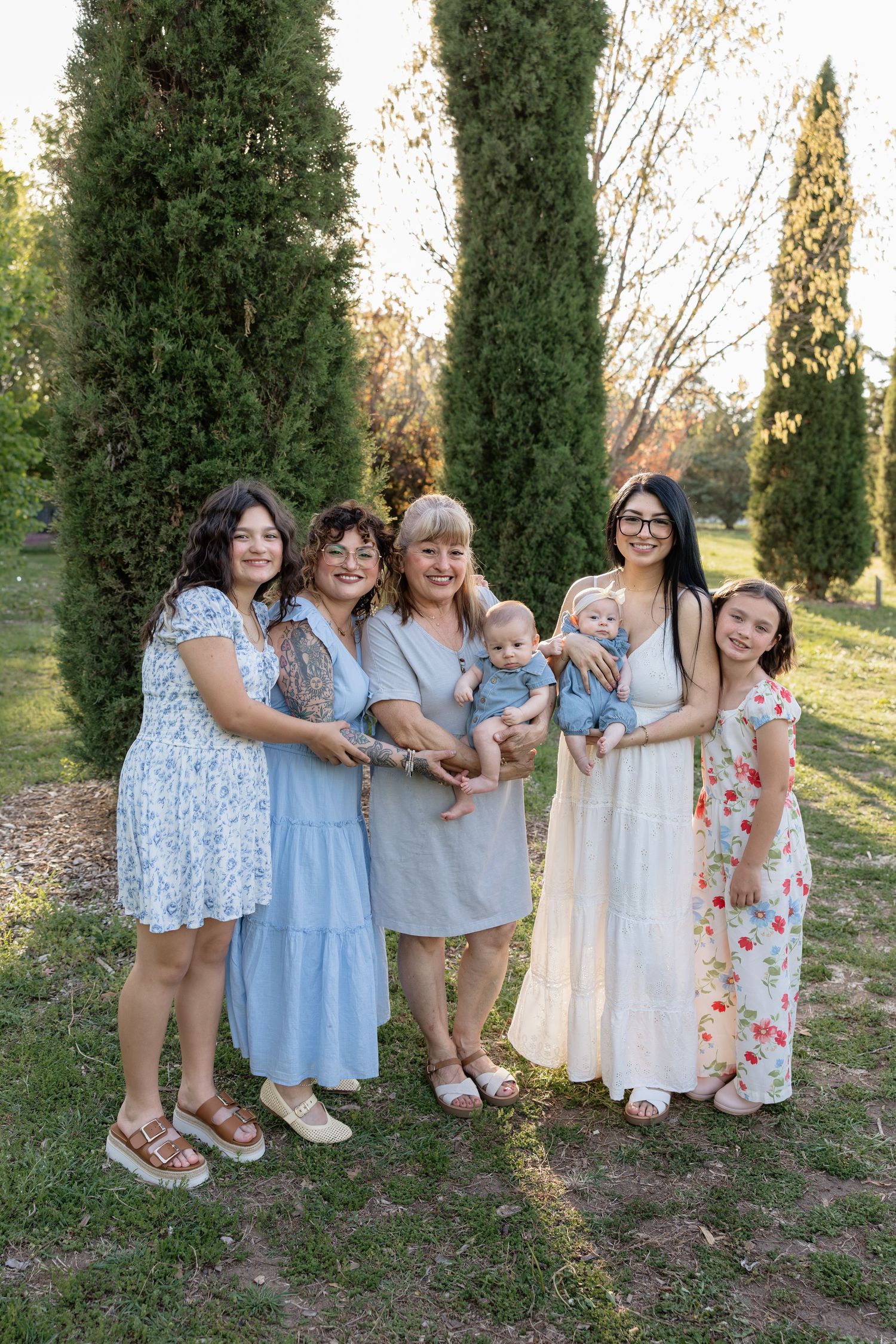 A multigenerational group of women and girls pose together outdoors holding a baby near tall cypress trees.